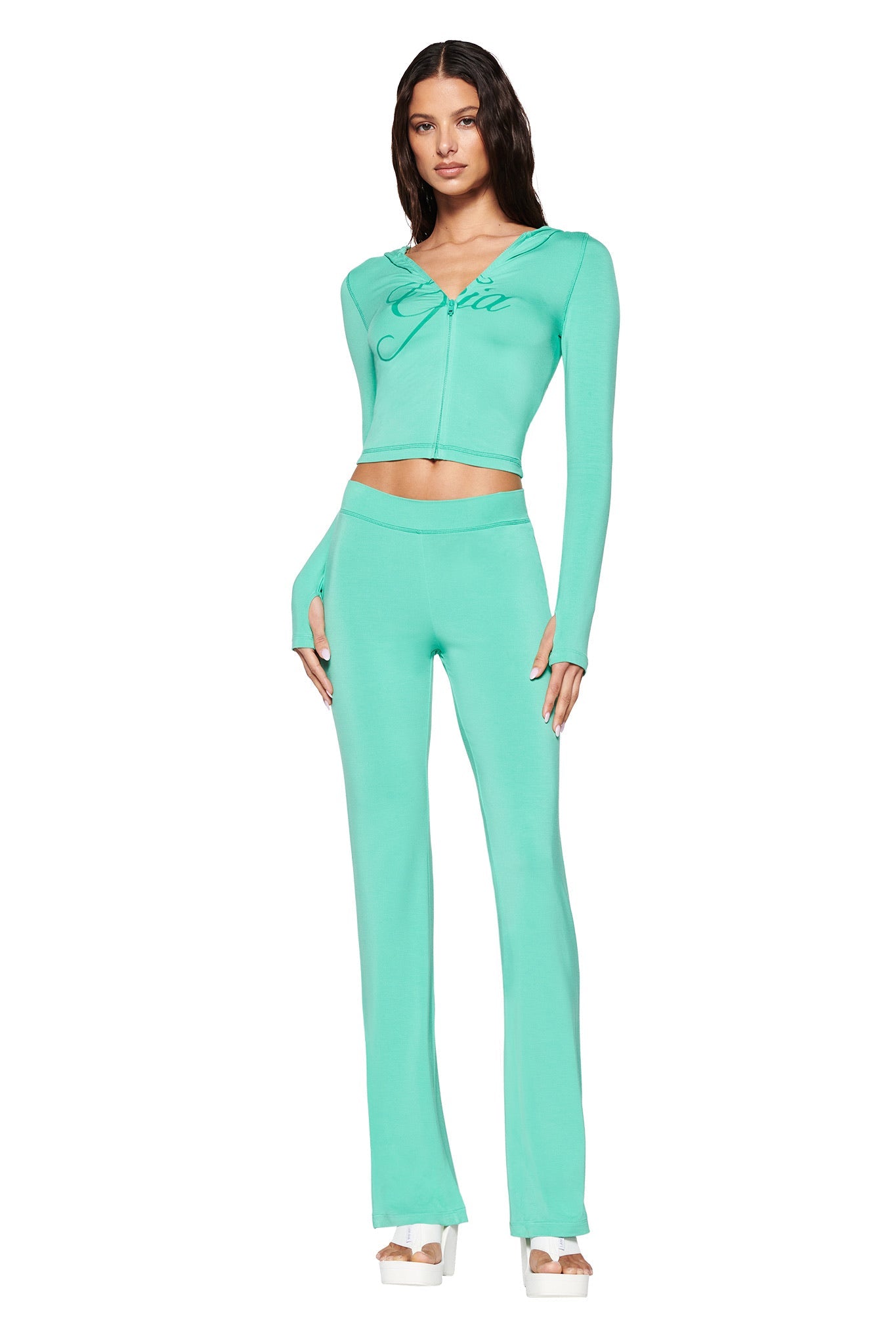 A woman models the I.AM.GIA Blare Trackpant in turquoise teal and a matching zip-up jacket, crafted from stretchy bamboo. The outfit complements her white platform sandals and long, wavy hair against a plain white background.