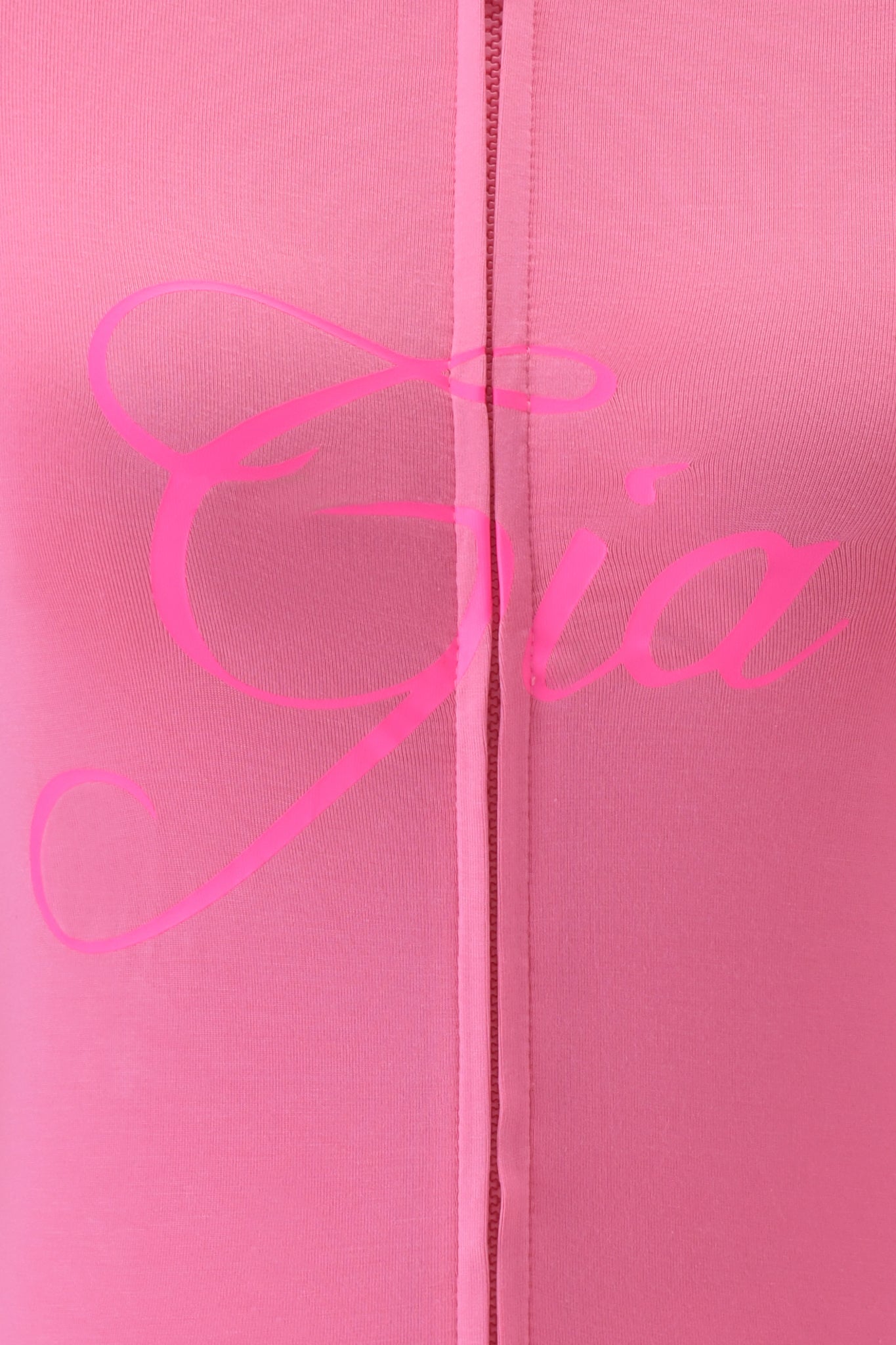 Close-up of the I.AM.GIA BLARE HOODIE in neon pink, showing its fabric with a vertical center seam and "Gia" in large, cursive, pink lettering across the material.