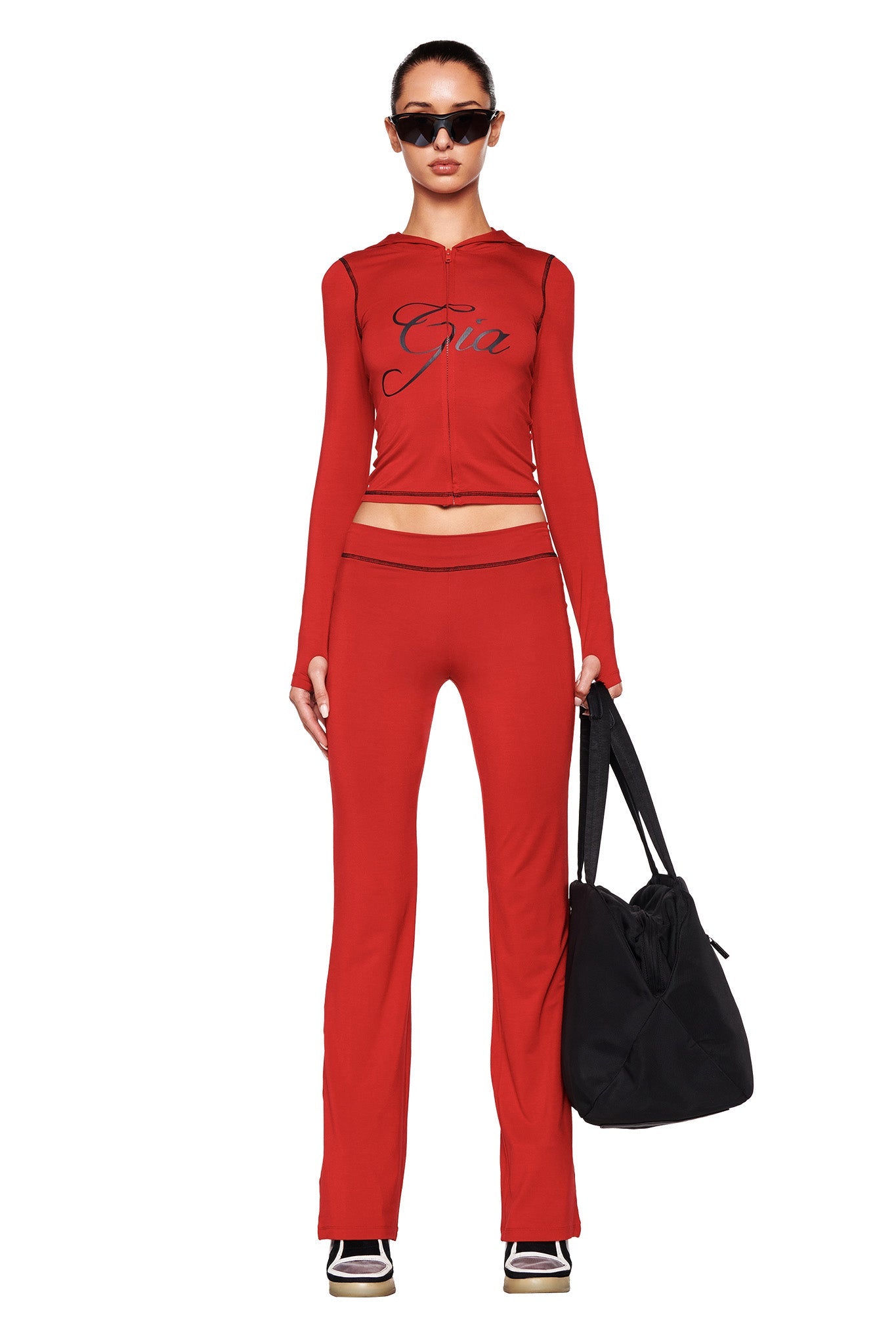 A person wearing the I.AM.GIA BLARE HOODIE in red and matching red pants stands against a white background. They are accessorized with black sunglasses and hold a large black bag in one hand.
