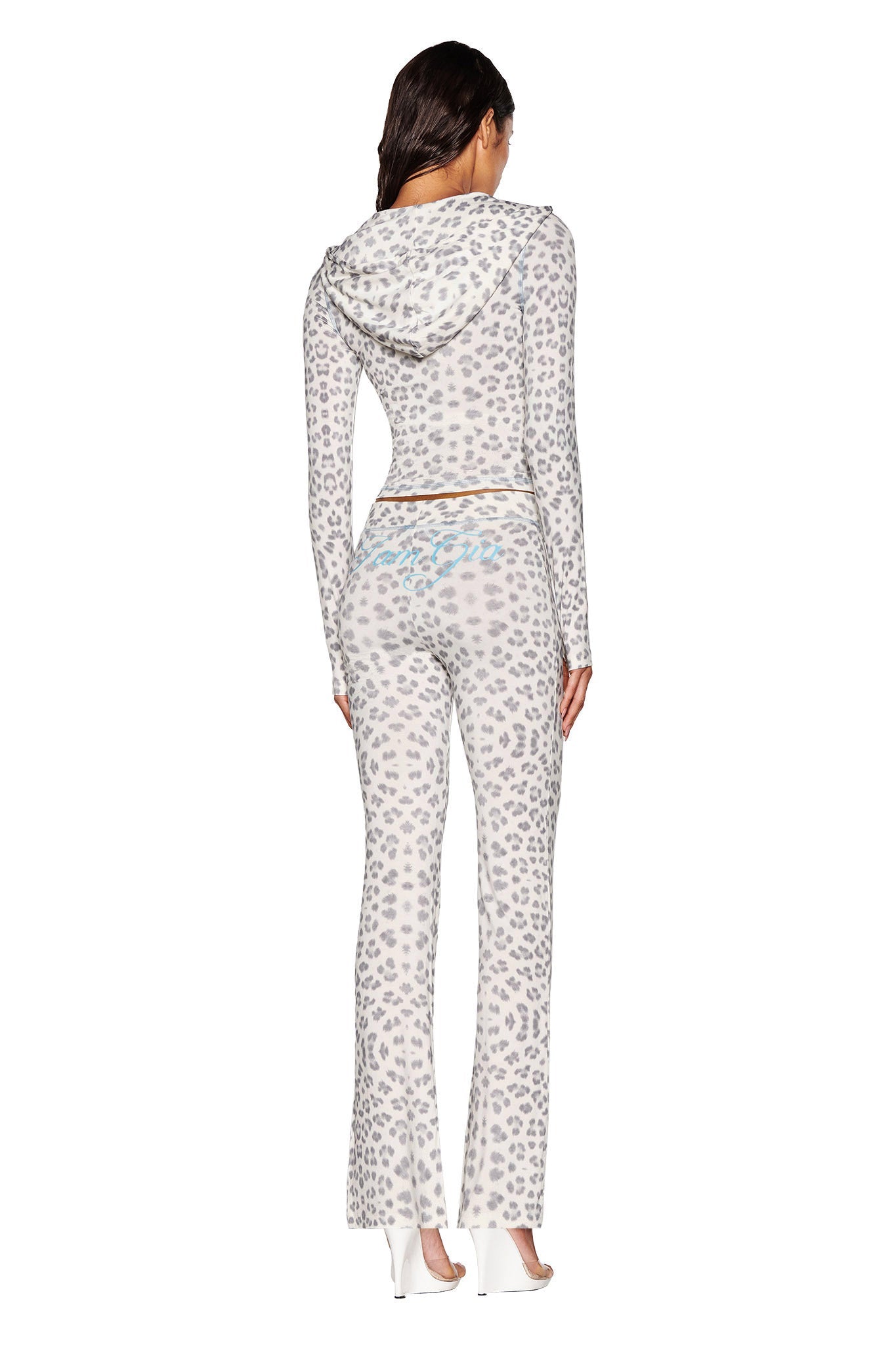 A person is standing with their back to the camera, wearing an I.AM.GIA BLARE TRACKPANT in Snow Leopard from the Winter Wild Card collection. The outfit highlights flared pants paired with white high heels against a plain white background.