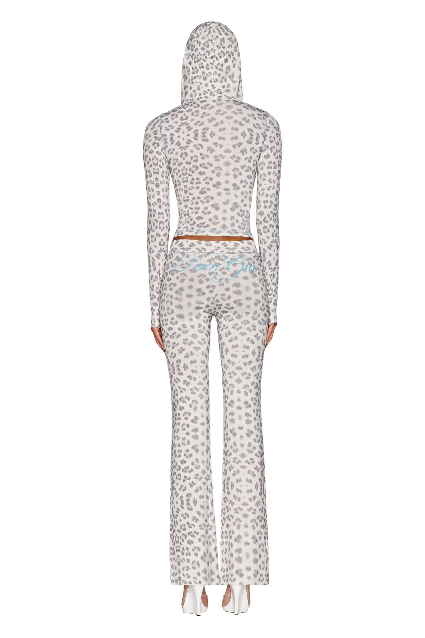 A person stands facing away, dressed in the I.AM.GIA BLARE HOODIE - SNOW LEOPARD from the WINTER WILD CARD collection, paired with matching bodycon fit pants. The outfit is complemented by sleek white high-heeled shoes, with a minimalist background that enhances its stylish impact against the plain white setting.