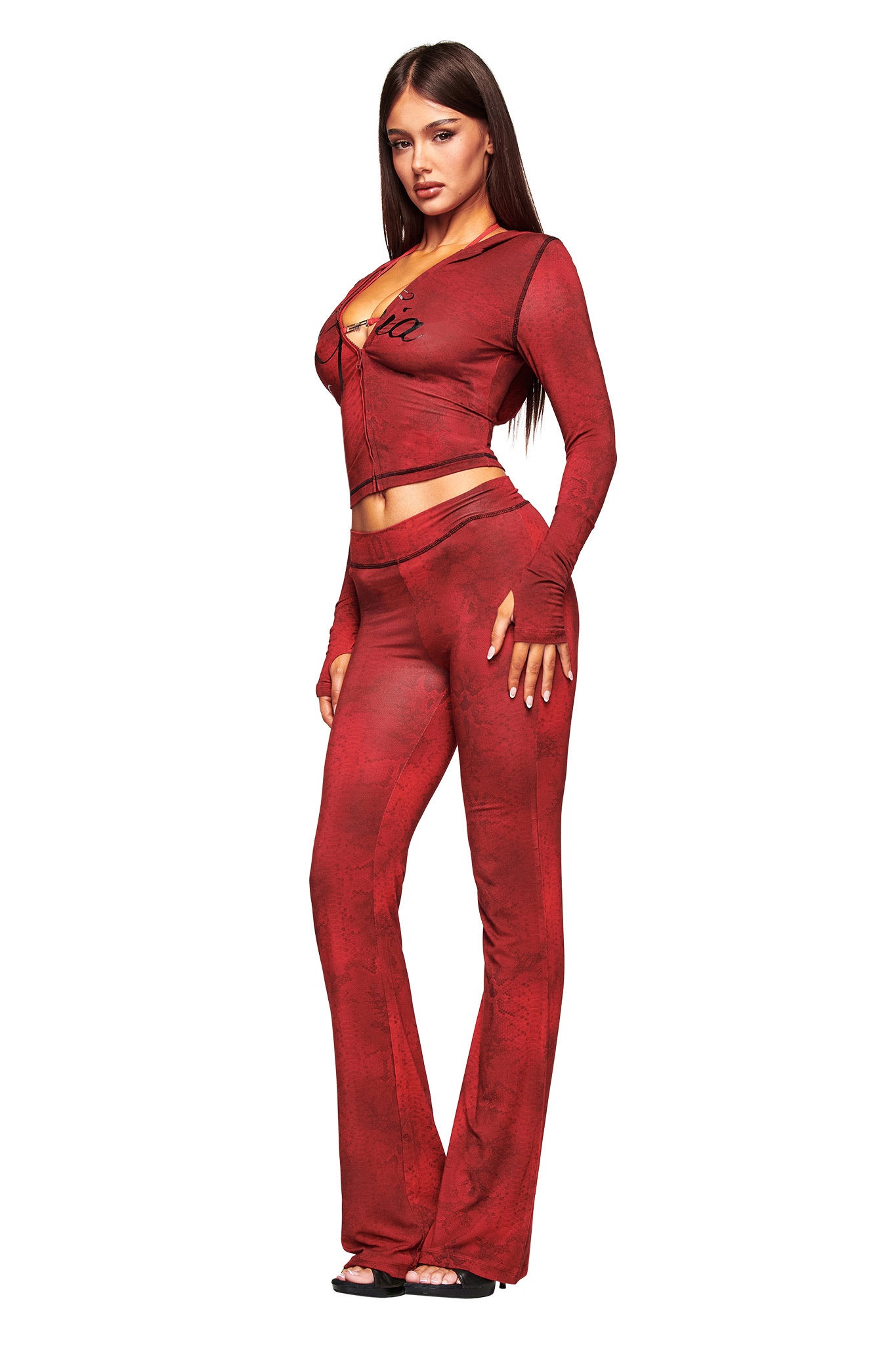 A woman poses against a white background wearing I.AM.GIA’s BLARE TRACKPANT in red serpentine, paired with a matching fitted zip-up top. She has long straight hair, neutral makeup, and black open-toe heels.