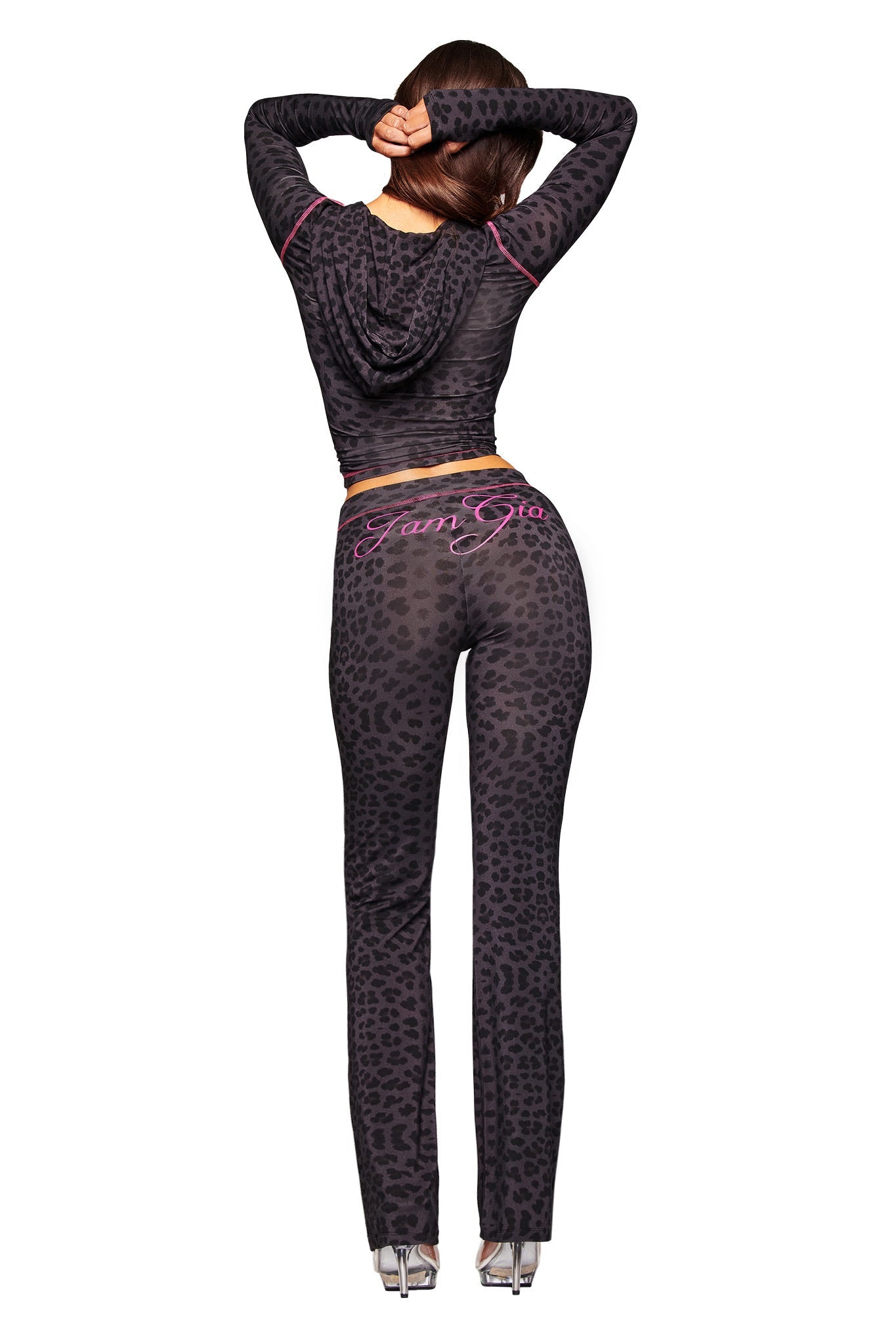 A woman faces away from the camera in a fitted, dark leopard-print I.AM.GIA BLARE HOODIE - PANTHER and matching pants featuring pink "FamGia" lettering on the back, finishing the outfit with clear high heels.