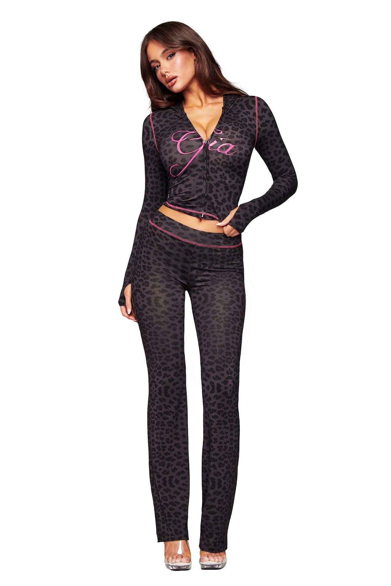 A woman poses in fitted, long-sleeve panther print top and matching I.AM.GIA BLARE TRACKPANT. Her top features "Gia" in pink script. She wears clear heels and stands against a white background.