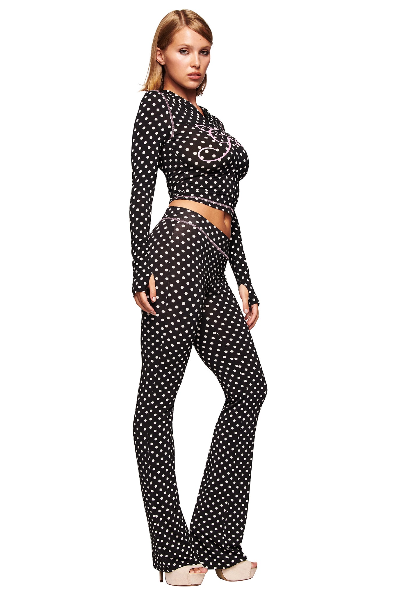 A woman poses against a white background wearing the I.AM.GIA BLARE HOODIE - BLACK POLKA DOT with matching black sheer long-sleeve top and flared pants, all featuring a white polka dot pattern, styled with high-heeled shoes.
