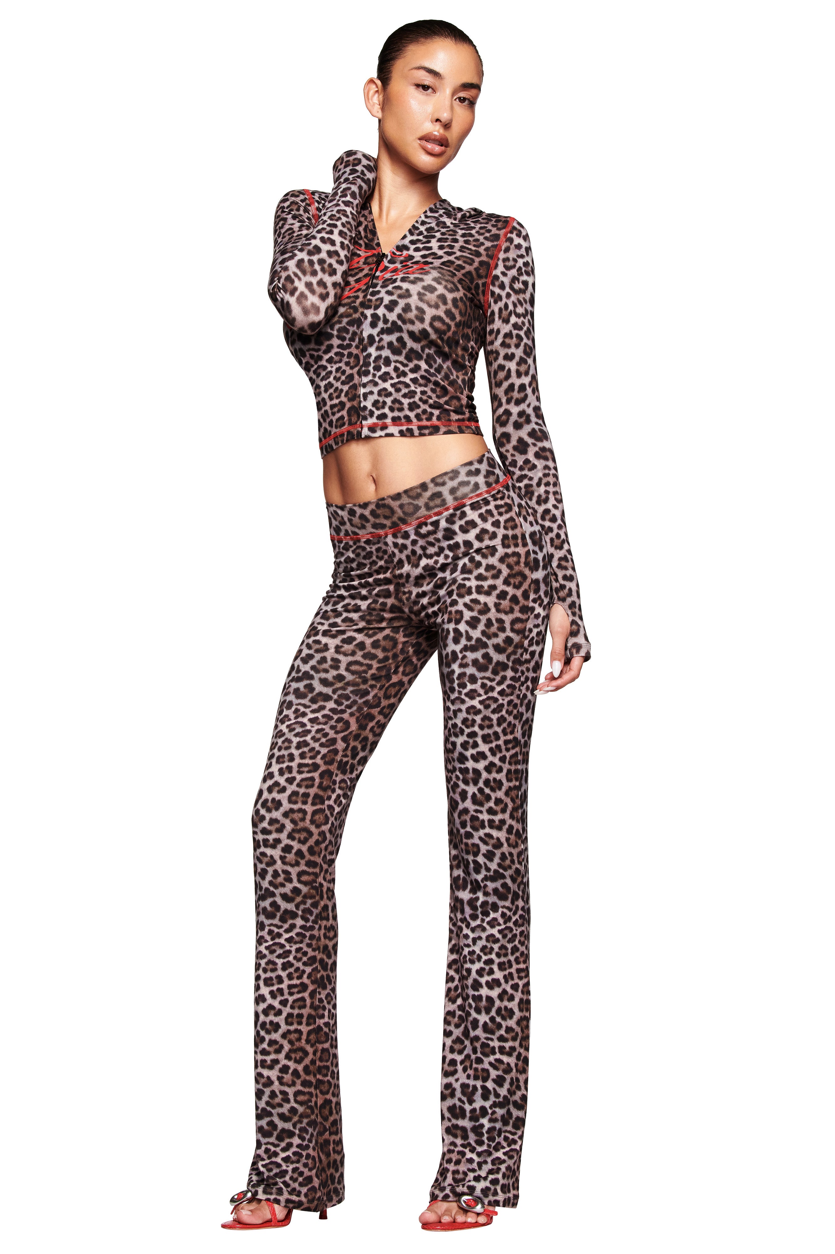A person poses in a fitted, long-sleeve dark leopard print top and matching BLARE TRACKPANT - DARK LEOPARD by I.AM.GIA, featuring a low-rise waist and red trim, styled with red open-toe heels and slicked-back hair for a bold look.