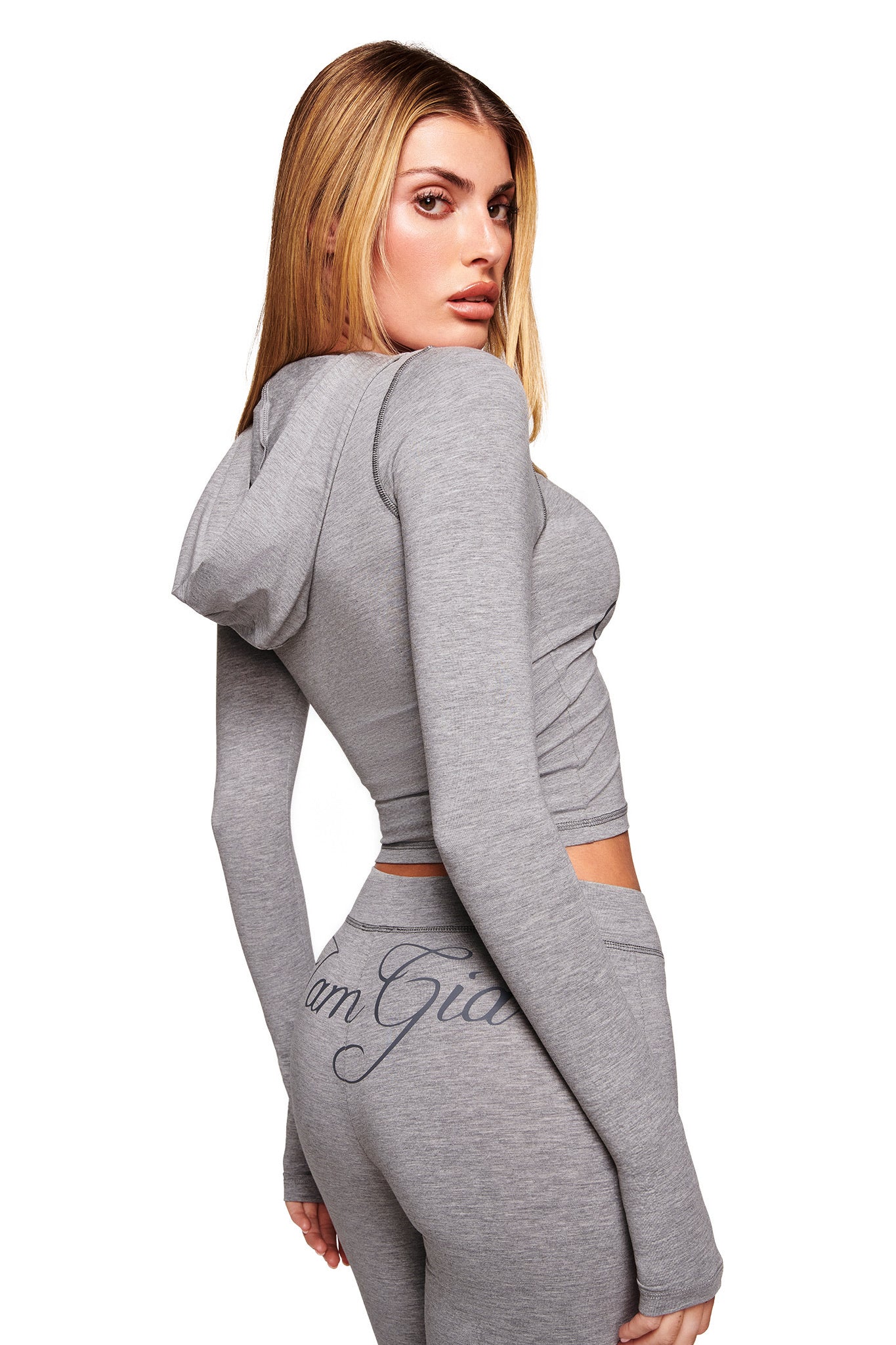 A woman with long blonde hair models I.AM.GIA’s BLARE JERSEY TRACKPANT in heather grey with a flared leg and logo on the back, paired with a fitted hoodie, posing against a white background.