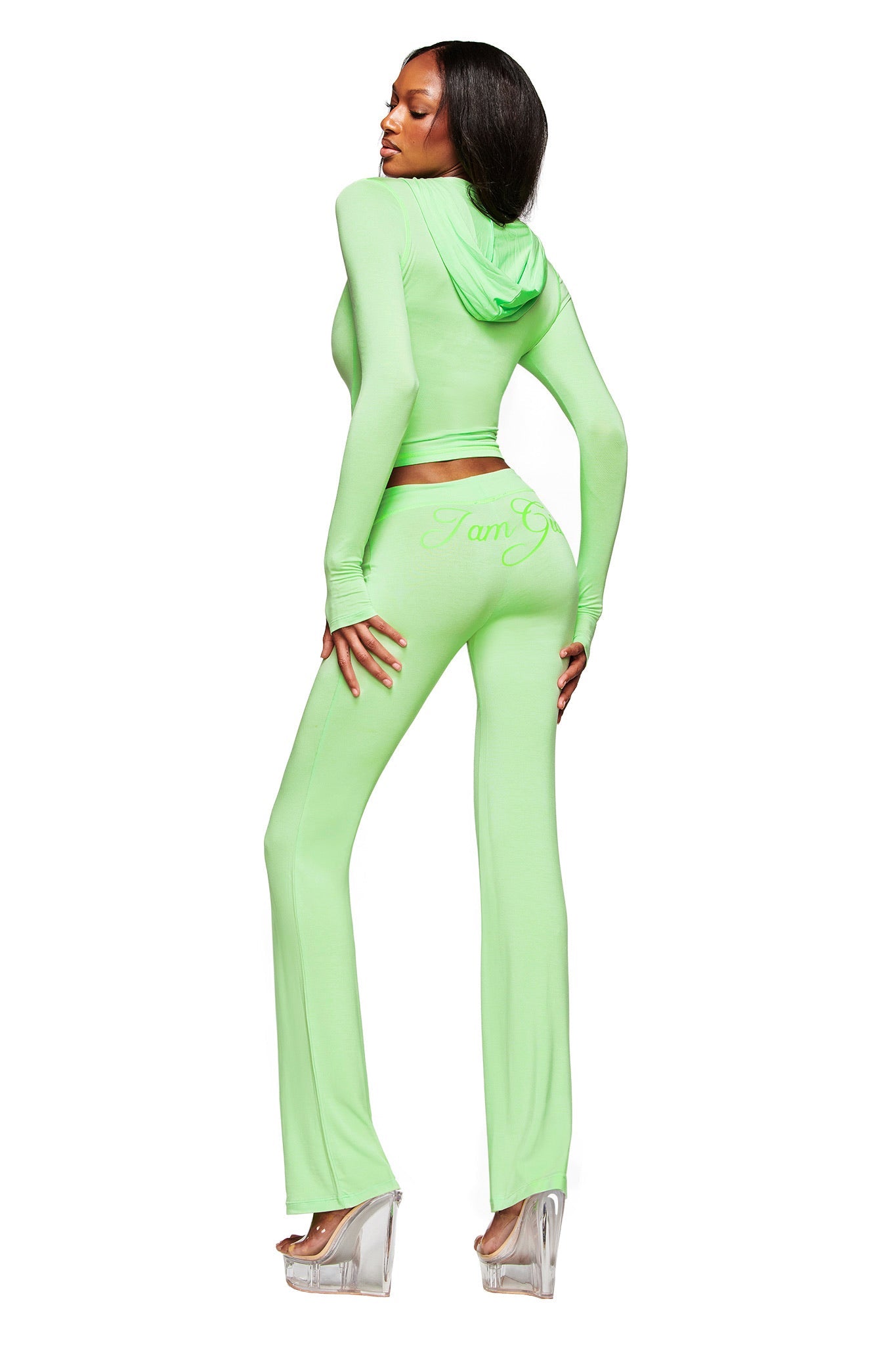 A woman models the I.AM.GIA BLARE HOODIE in neon green with matching flared pants, featuring the "I Am G" slogan on the back. She faces away, looking over her shoulder in clear platform heels against a white background.