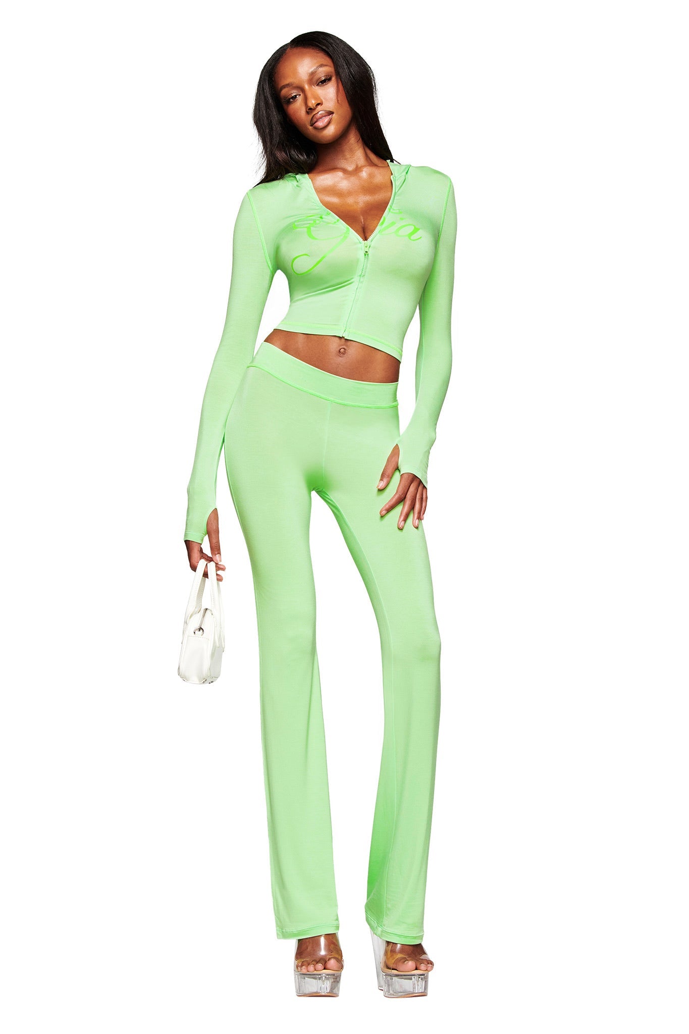 A woman poses in the I.AM.GIA BLARE HOODIE in neon green with matching high-waisted pants, subtle GIA logo, silver platform heels, and a small white handbag against a white background.