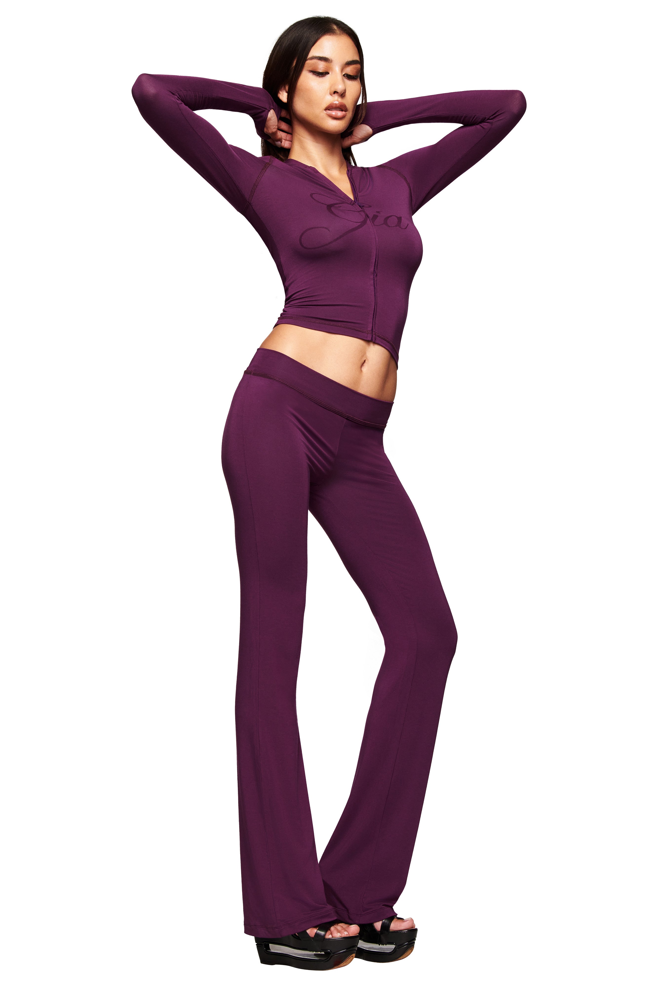A woman poses confidently in I.AM.GIA's BLARE TRACKPANT - PLUM, styled with a fitted, long-sleeve purple zip-up top and black platform sandals. She stands against a white background, arms raised and eyes closed.
