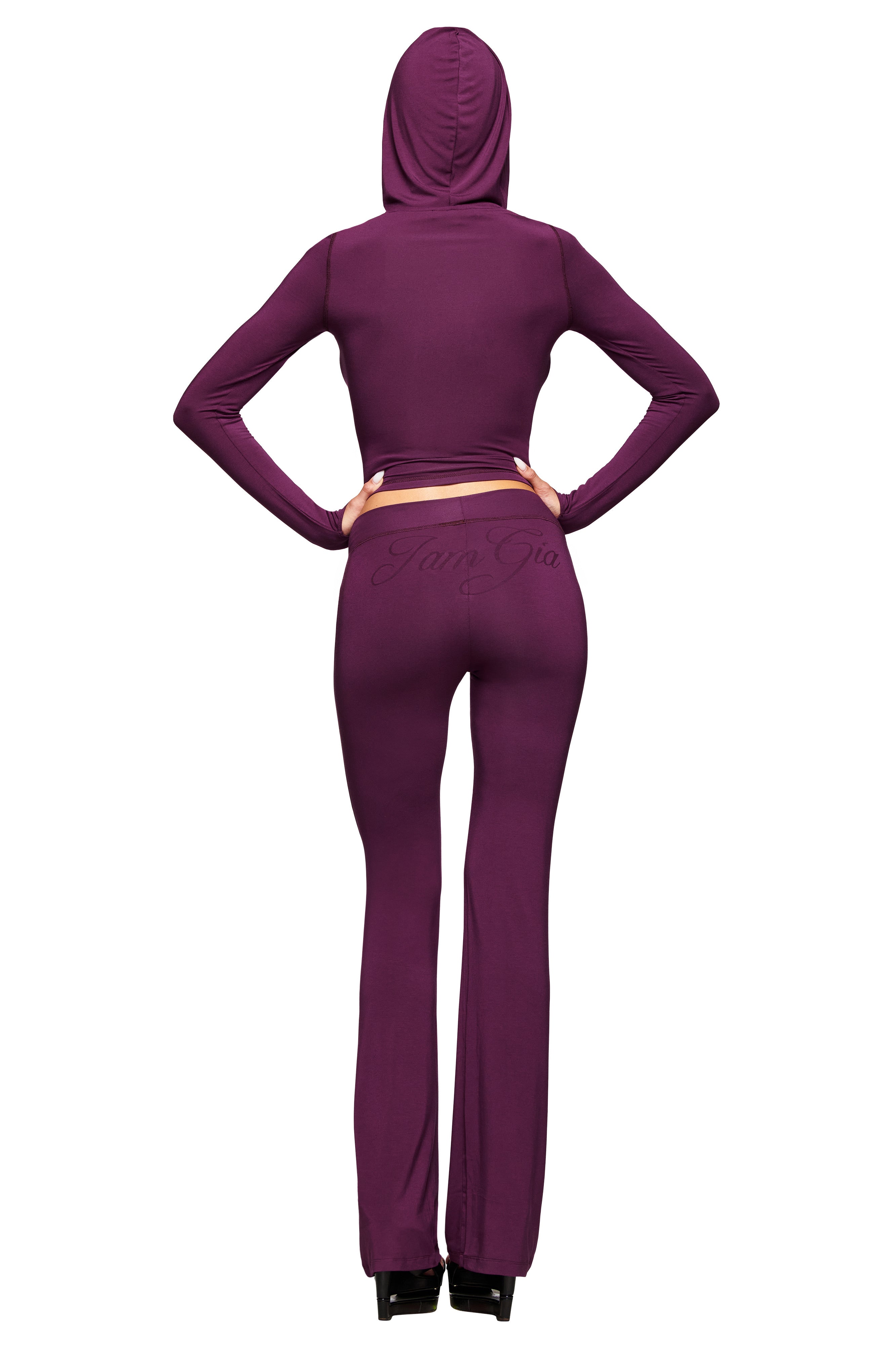 A person stands facing away, wearing the I.AM.GIA BLARE TRACKPANT in plum—a fitted, long-sleeve, hooded tracksuit with a low-rise waist, flared legs, and subtle text on the back. Hands on hips; paired with black shoes.