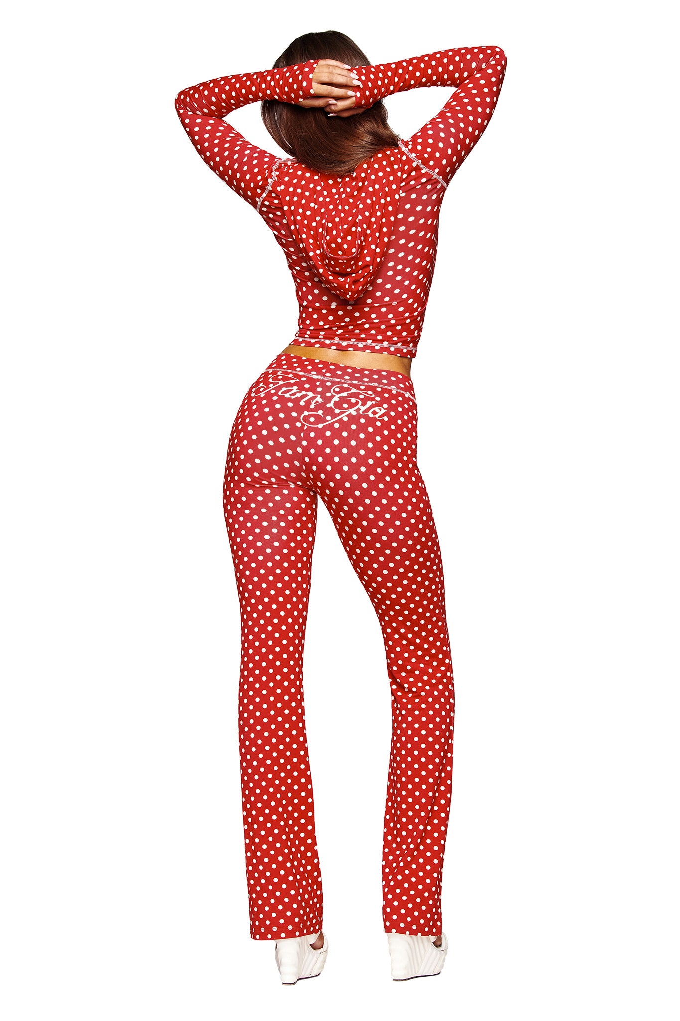 A person stands facing away, arms raised, wearing I.AM.GIA's BLARE TRACKPANT - RED POLKA DOT with a matching long-sleeve hooded top and white heels.