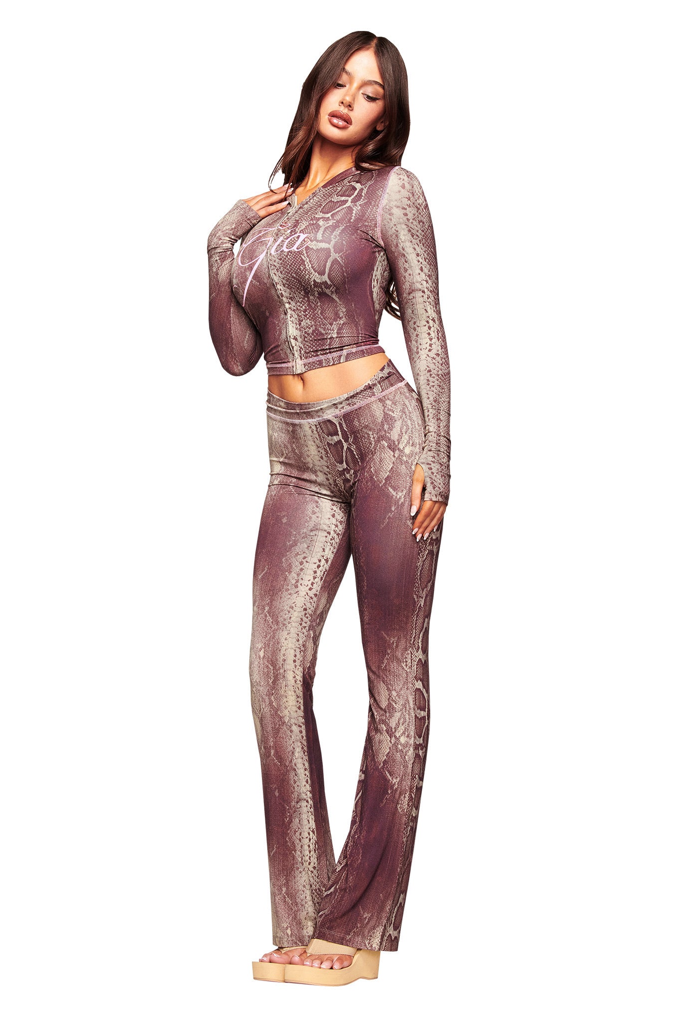 A woman in a mauve and taupe snake print crop top and matching high-waisted, wide-leg pants with beige platform sandals poses against a white background—perfectly paired with the I.AM.GIA BLARE HOODIE - SNAKE for cool evenings.