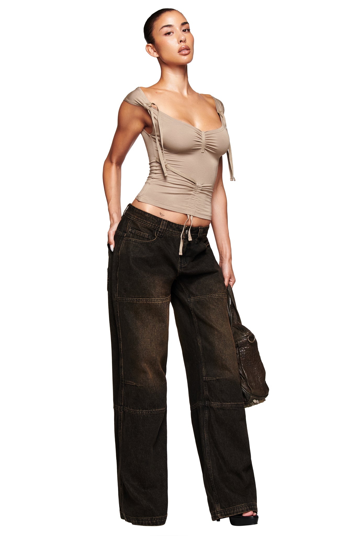 A woman poses confidently against a plain white background wearing I.AM.GIA’s MISTY JEAN - BLACK, paired with a beige ruched cap-sleeve top and holding a brown handbag by her side.