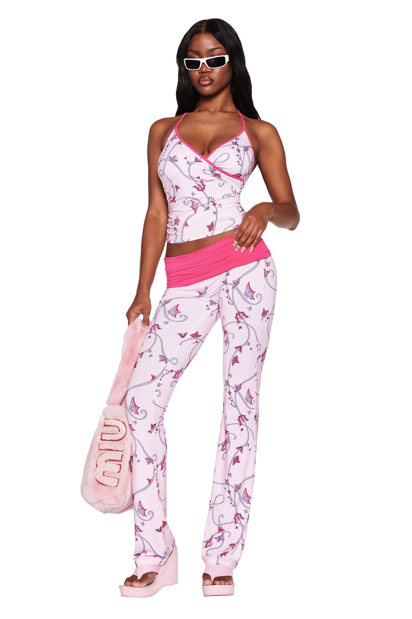 A person confidently wears the I.AM.GIA JANNIS TANK in pink with a butterfly ribbon, styled with platform sandals and sunglasses, holding a pink handbag against a white background.