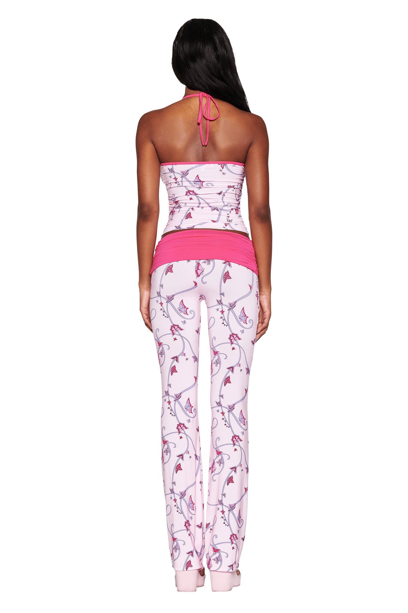 A person with long hair is seen from the rear wearing the JANNIS TANK - PINK : BUTTERFLY RIBBON by I.AM.GIA, which is a pink and white halter top with leggings. The bodycon fit emphasizes a floral pattern with pink accents and a butterfly ribbon print at the waistband, set against a plain white background.