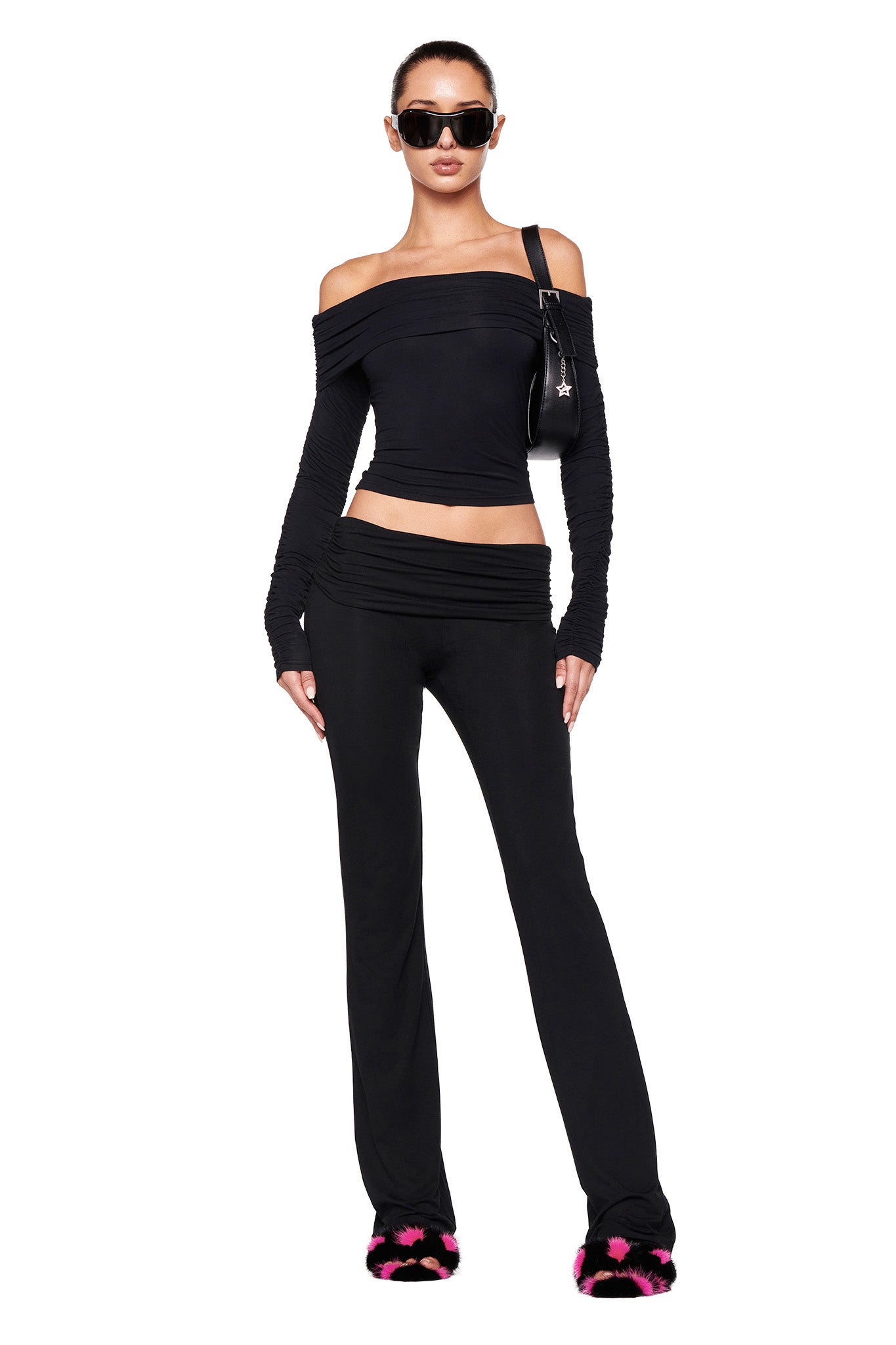 A woman stands confidently wearing an off-the-shoulder black top paired with the JANNIS PANT in solid black from I.AM.GIA. She accessorizes with dark sunglasses, a small black shoulder bag, and furry black slippers featuring pink accents. Her hair is pulled back neatly.