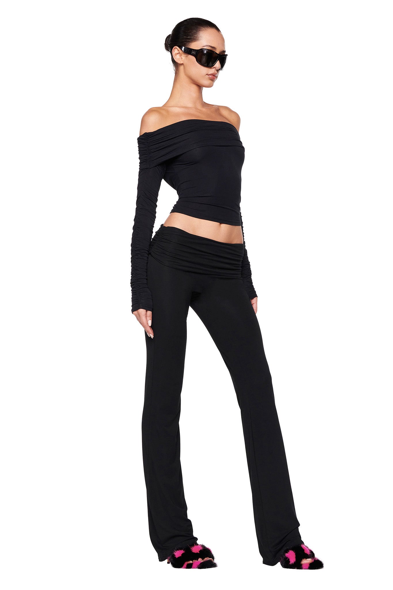 A person in a fashionable, all-black I.AM.GIA outfit poses against a white background. They are wearing an off-shoulder top, the JANNIS PANT - BLACK: SOLID BLACK high-waisted pants, and black sunglasses. The individual also has their hair tied back in a bun and is wearing black slippers with pink accents.