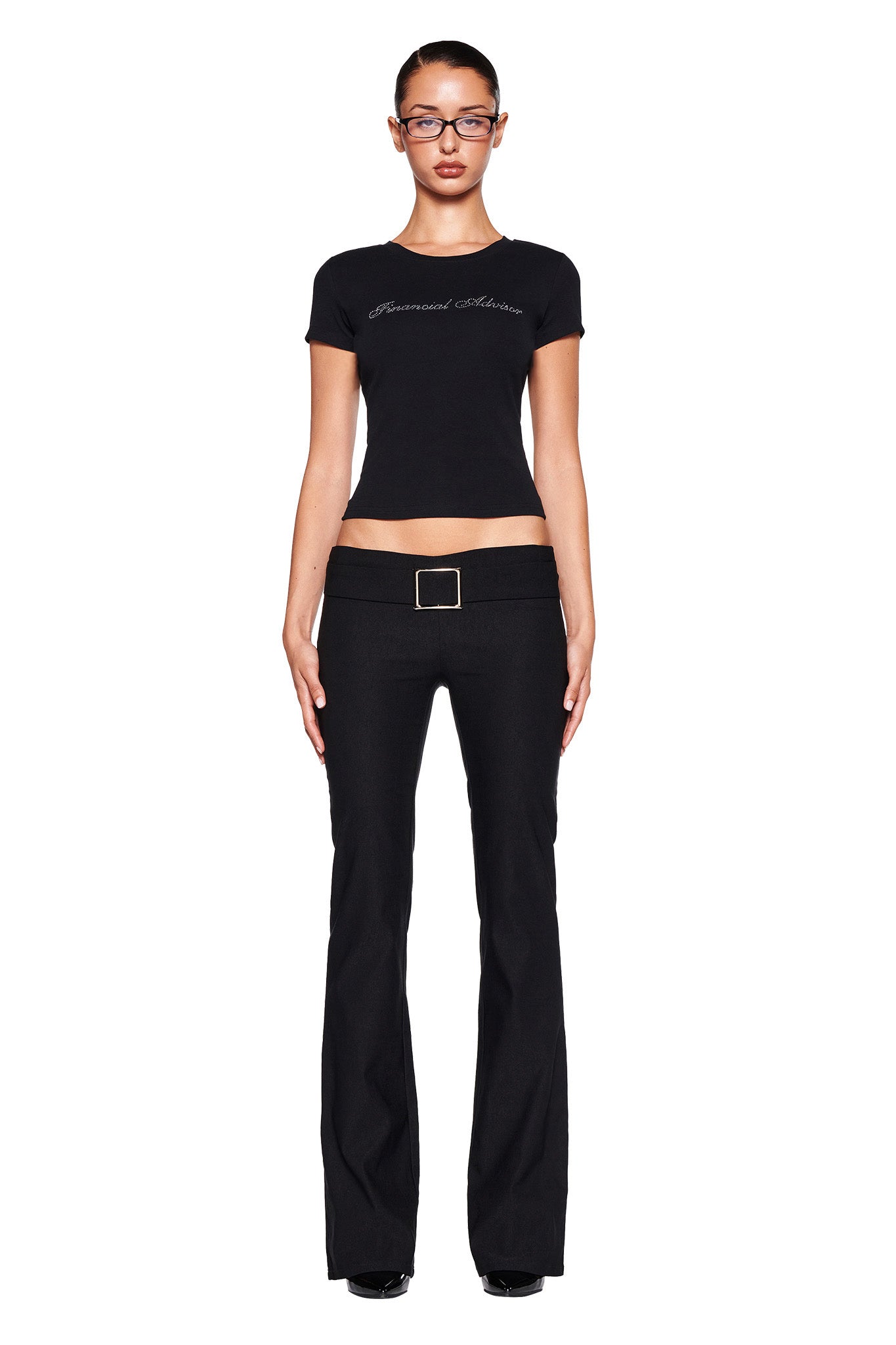 A person stands against a white background, sporting a fitted black t-shirt with text across the chest and the sleek SHARAH PANT - BLACK from I.AM.GIA, which feature a large rectangular belt buckle. With their hair tied back and glasses on, they complete the look effortlessly.