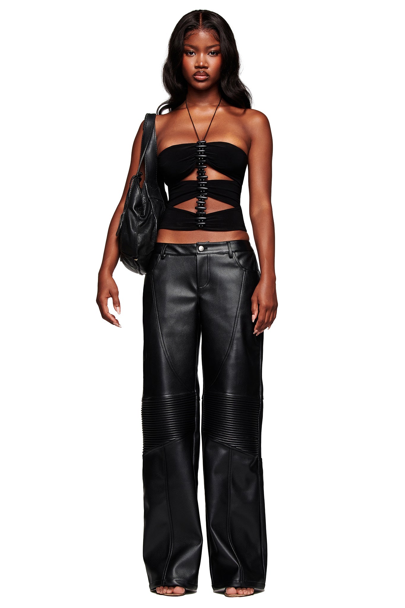 A woman with long, wavy hair and a neutral expression stands facing forward wearing the KRISTIE PANT - BLACK by I.AM.GIA, paired with a black strapless cut-out top and holding a black shoulder bag.