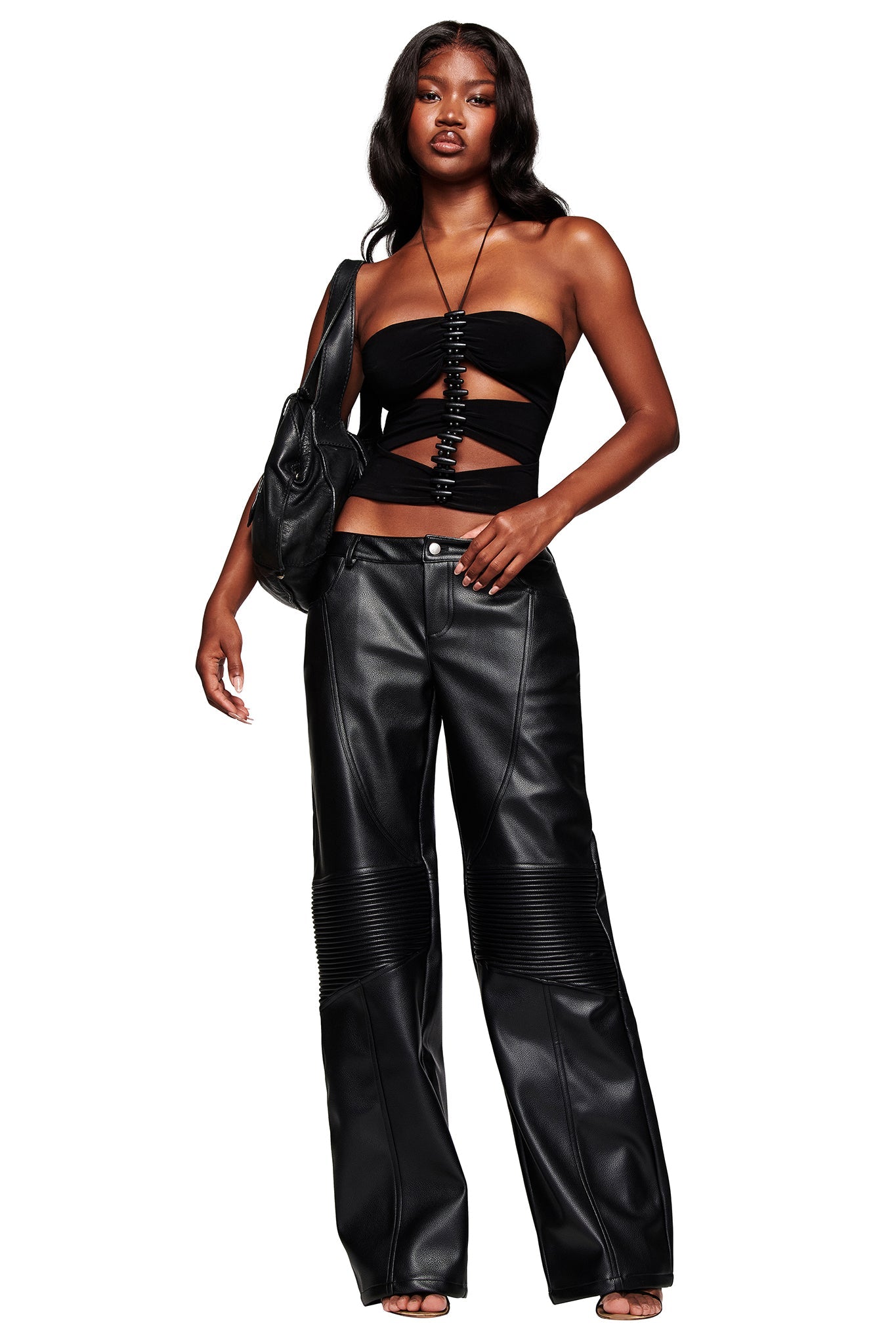 A woman poses against a white background wearing the I.AM.GIA KRISTIE PANT in black, styled with a strapless cut-out top, heeled sandals, a necklace, and a black shoulder bag for a chic look.