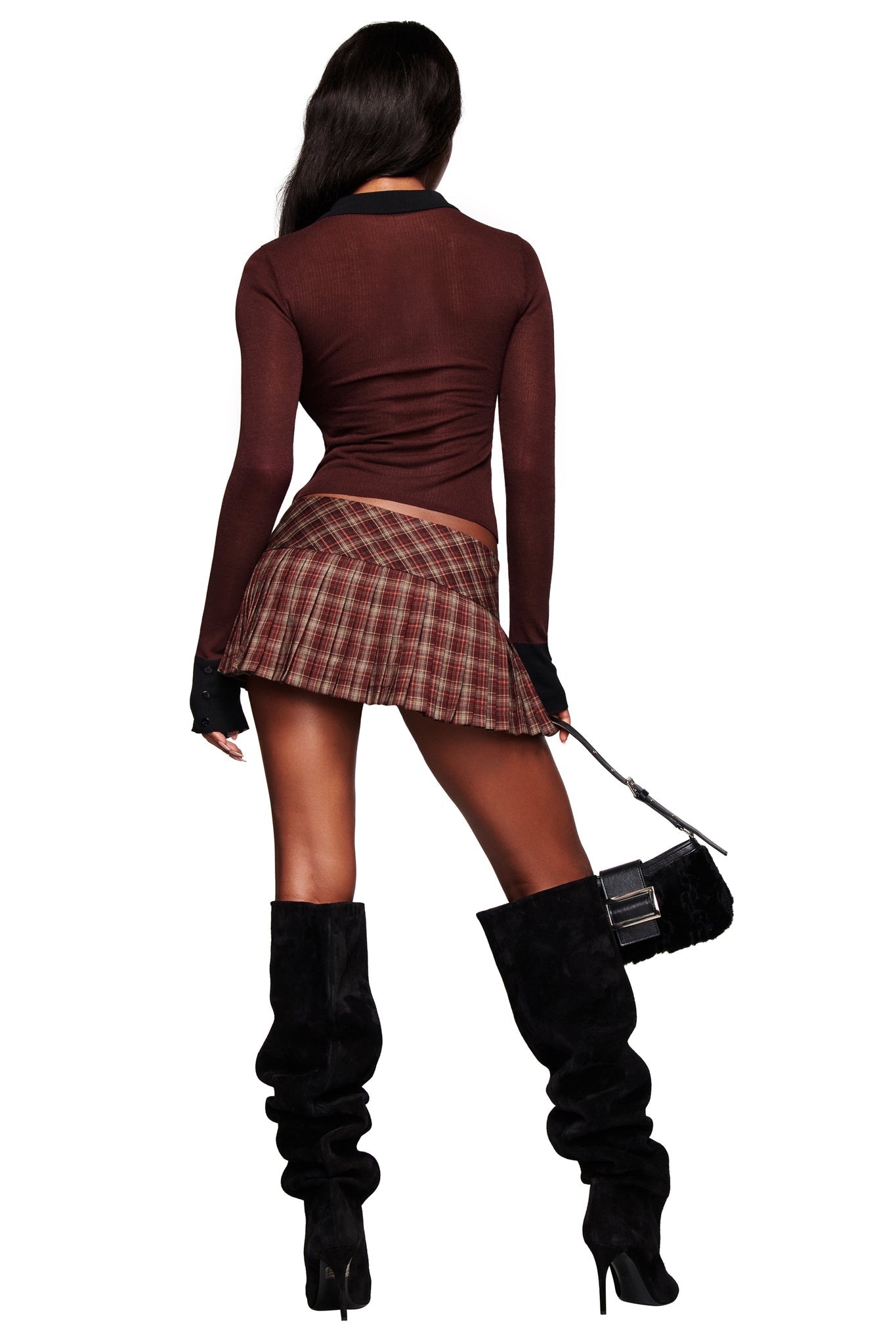 A woman faces away, wearing the I.AM.GIA DAVINA TOP in brown, a short plaid skirt, tall black slouchy boots, and holding a small black handbag. Her long hair flows loosely down her back.