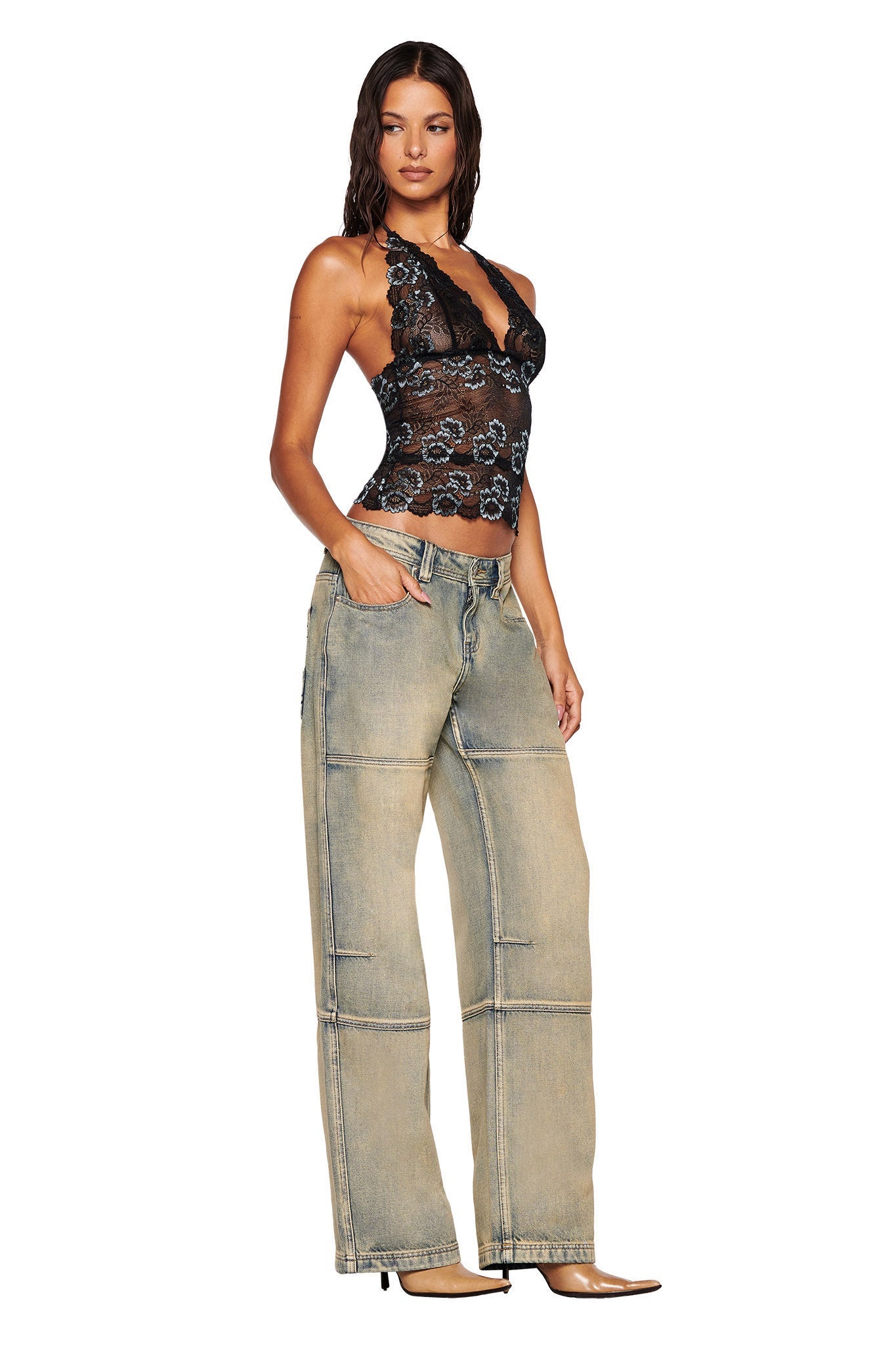 In a studio setting, a woman confidently poses wearing the GEM HALTER TOP in black from I.AM.GIA, paired with baggy beige jeans. Her long brown hair cascades down as she stands with one hand in her pocket and the other resting by her side, capturing the camera's attention.