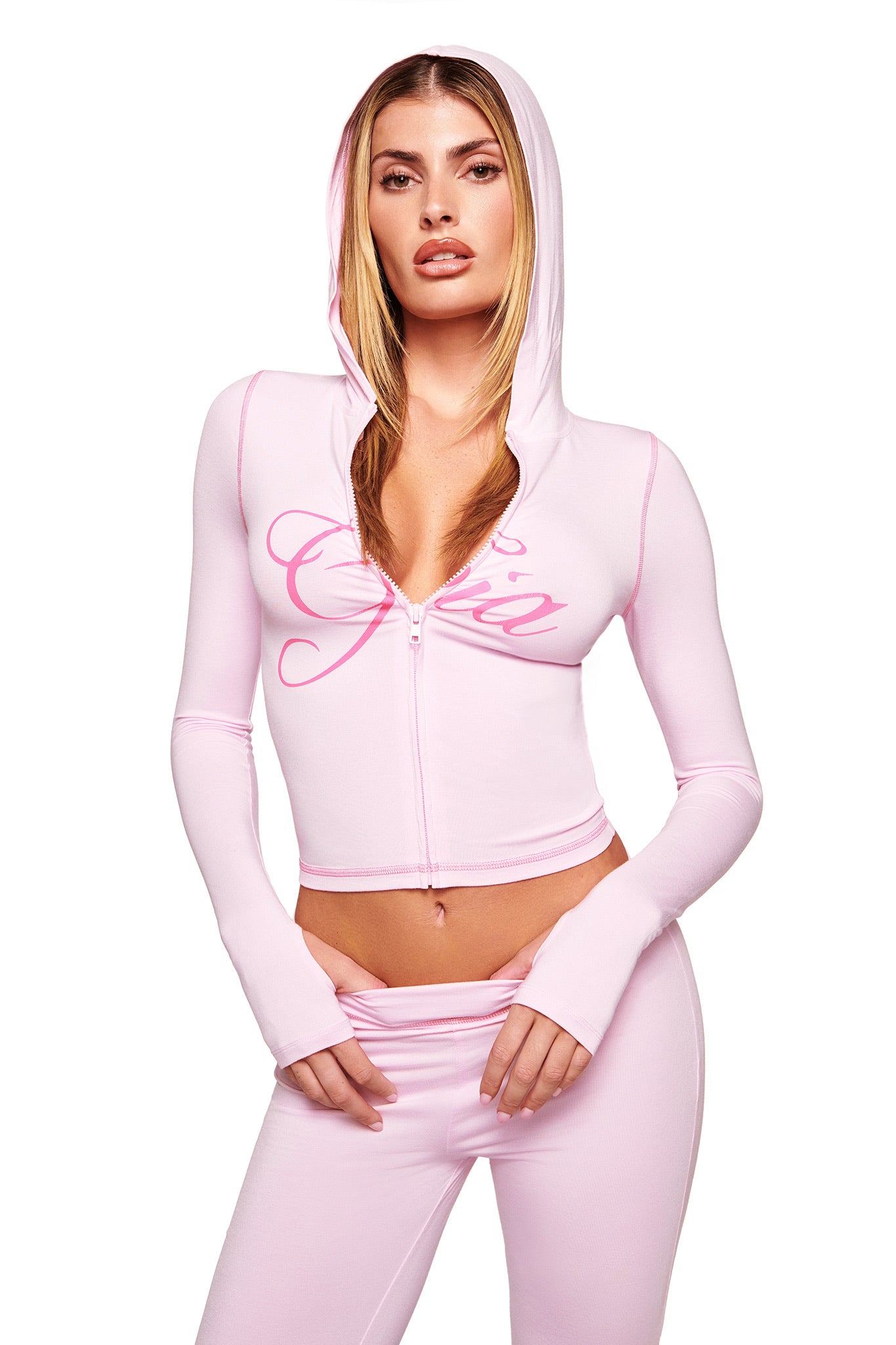 A woman poses confidently against a white background, wearing I.AM.GIA’s pastel pink BLARE TRACKPANT and a matching fitted zip-up hoodie with "Gia" scripted across the chest.