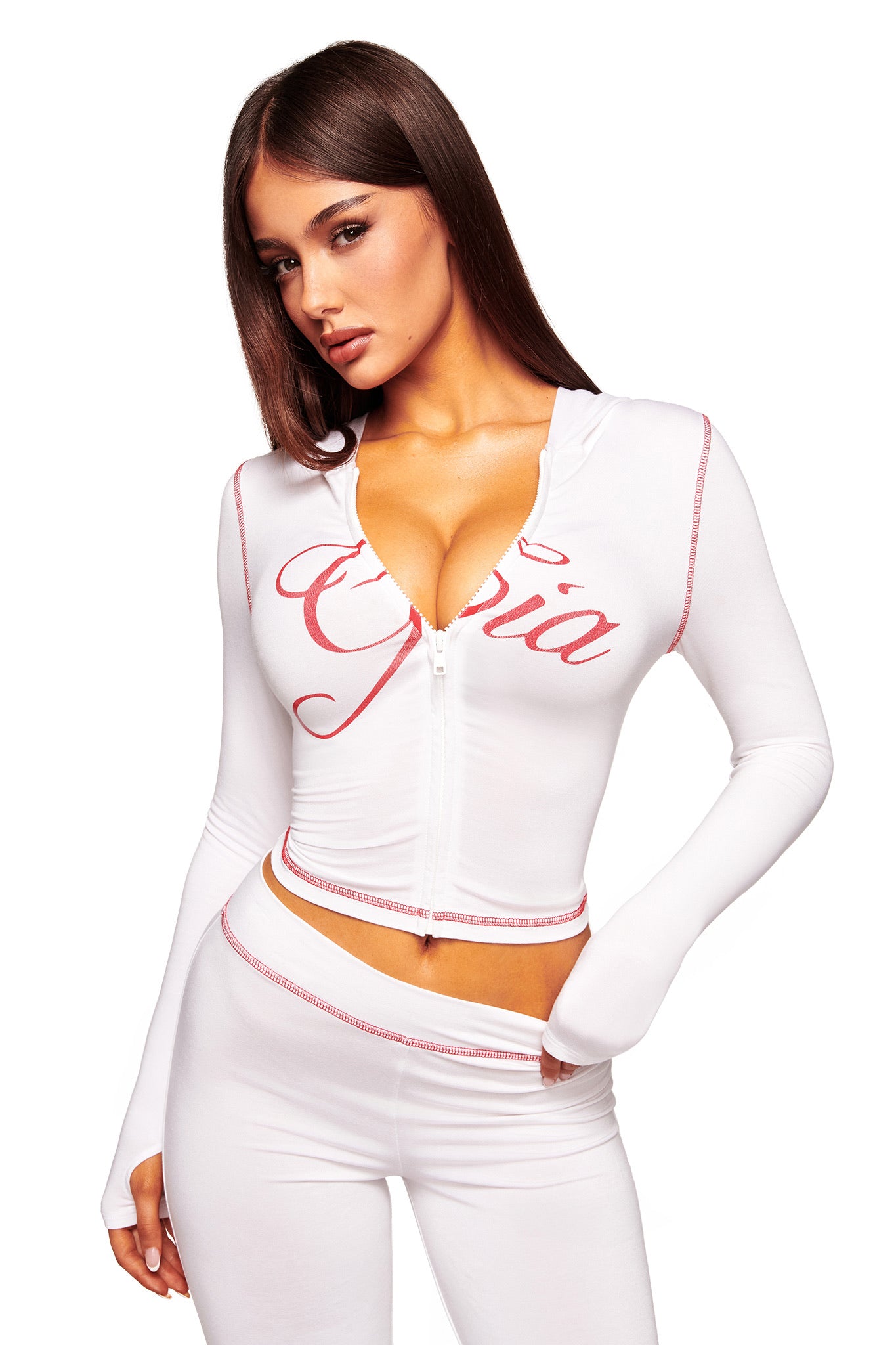 A woman with long brown hair models the I.AM.GIA BLARE HOODIE in white, featuring pink lettering, paired with matching white pants. She poses with one hand on her waist against a white background.