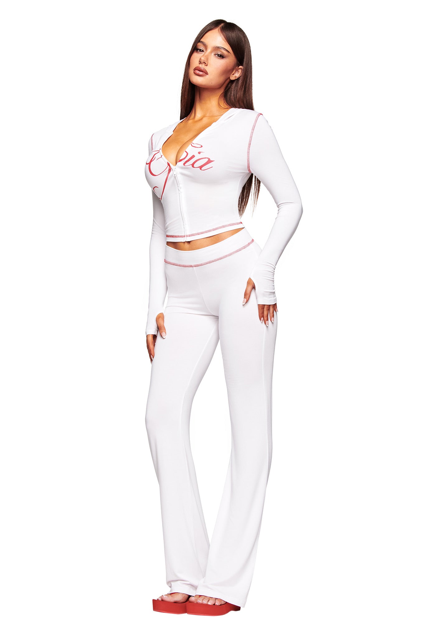 A woman poses confidently against a plain white background, wearing the I.AM.GIA BLARE HOODIE in white with matching high-waisted pants and red sandals, channeling signature I.AM.GIA style.