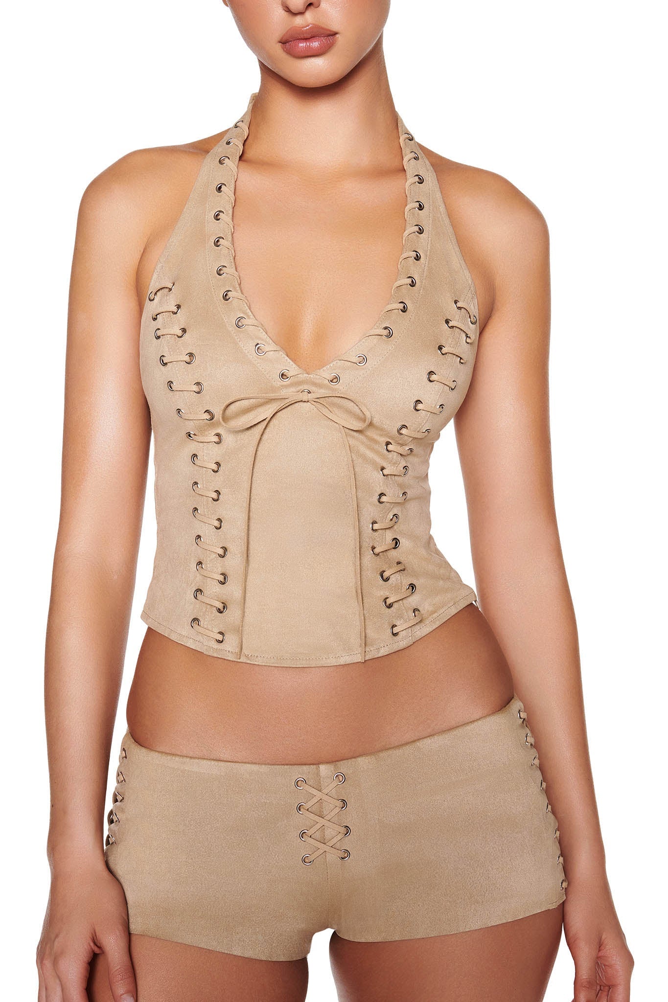 A person wearing I.AM.GIA's RAMONA SHORT - BEIGE : TAN, a faux suede short with lace-up details and silver eyelets, paired with a tan halter top featuring similar lace-up detailing and a center bow.