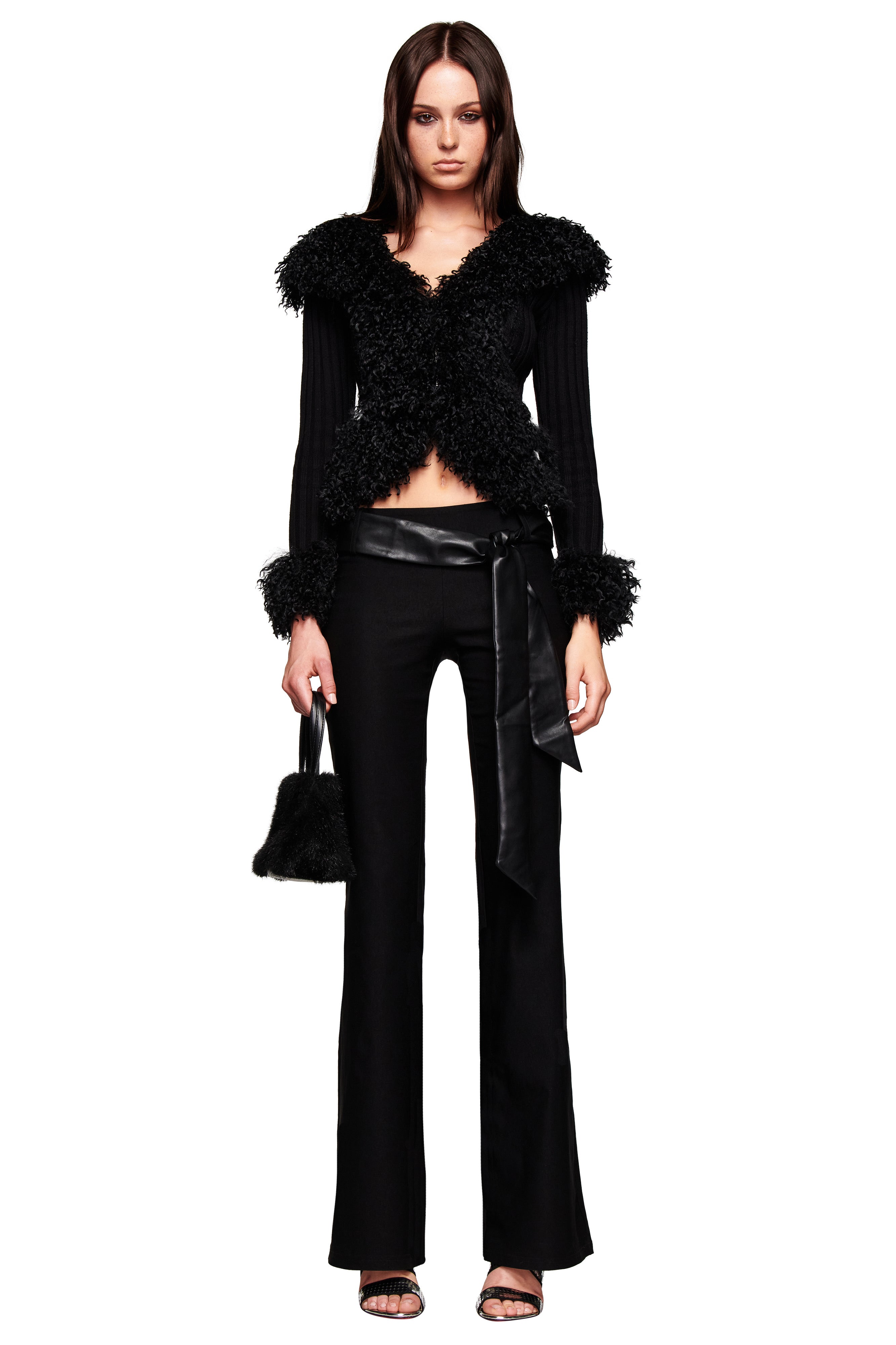 A woman with straight brown hair and a neutral expression wears the I.AM.GIA CRESSIDA JACKET - BLACK with a faux fur collar, flared black pants with a satin belt, black heeled sandals, and carries a small black fuzzy handbag.