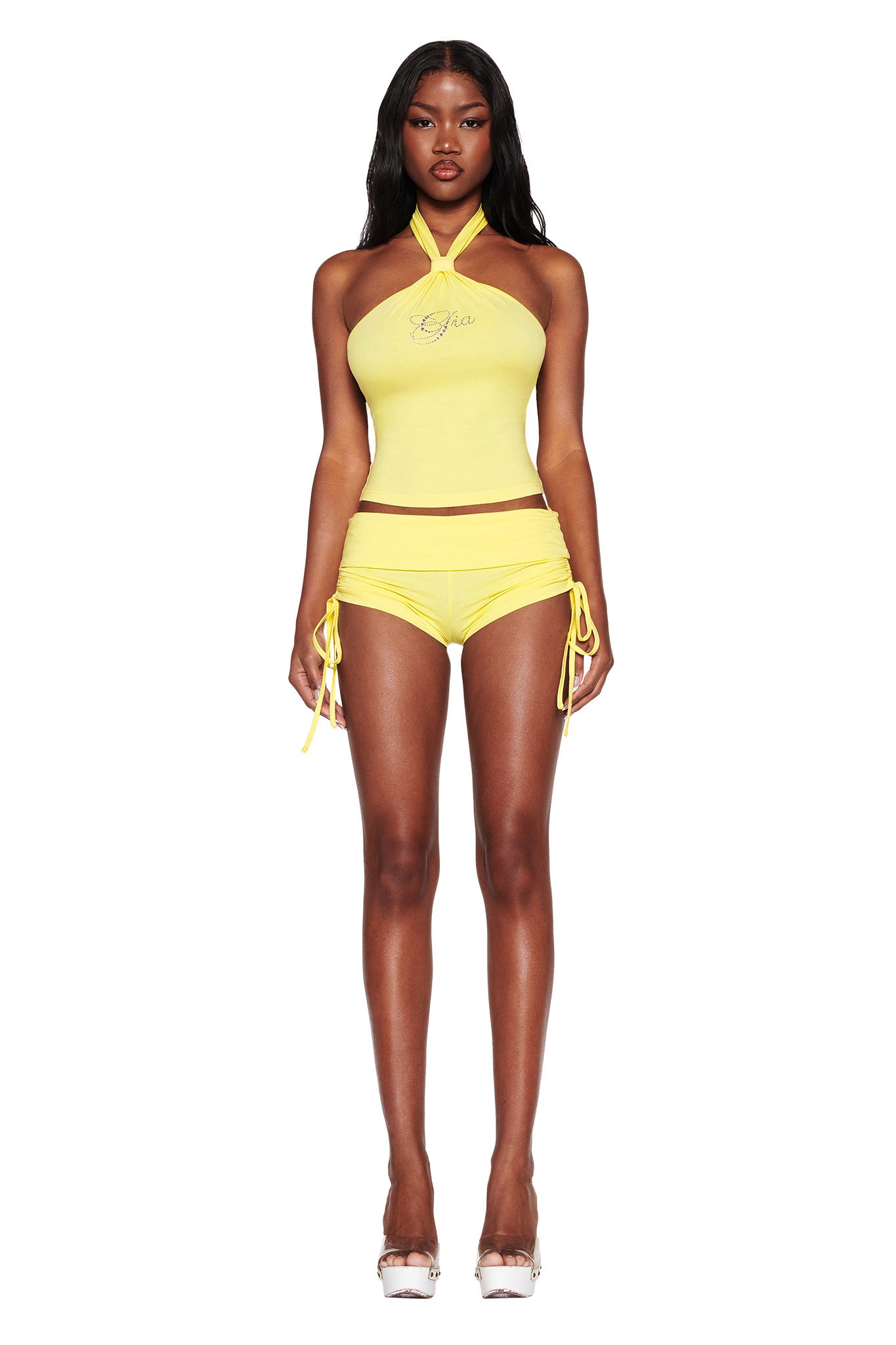 A person with long dark hair faces forward in a ROWAN TOP - YELLOW by I.AM.GIA, made from bamboo fabrication with text printed on it, paired with matching high-waisted shorts featuring side ties. They complement the outfit with white platform sandals.