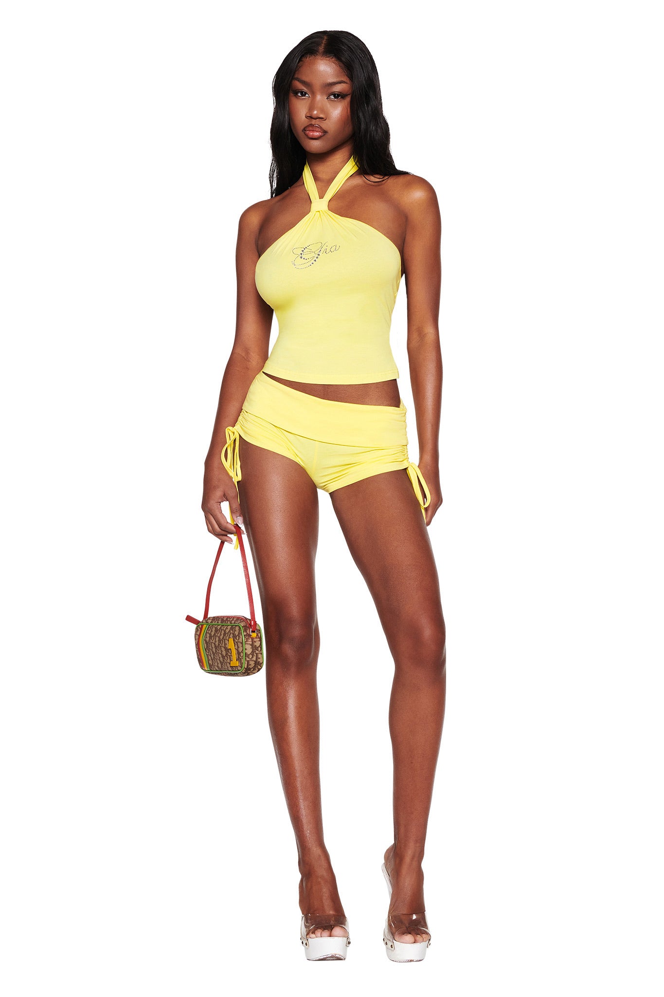 A person confidently wears the ROWAN TOP - YELLOW by I.AM.GIA with matching bodycon shorts, holding a small brown handbag. They have long black hair and wear clear high heels against a crisp white backdrop, highlighting their vibrant outfit.