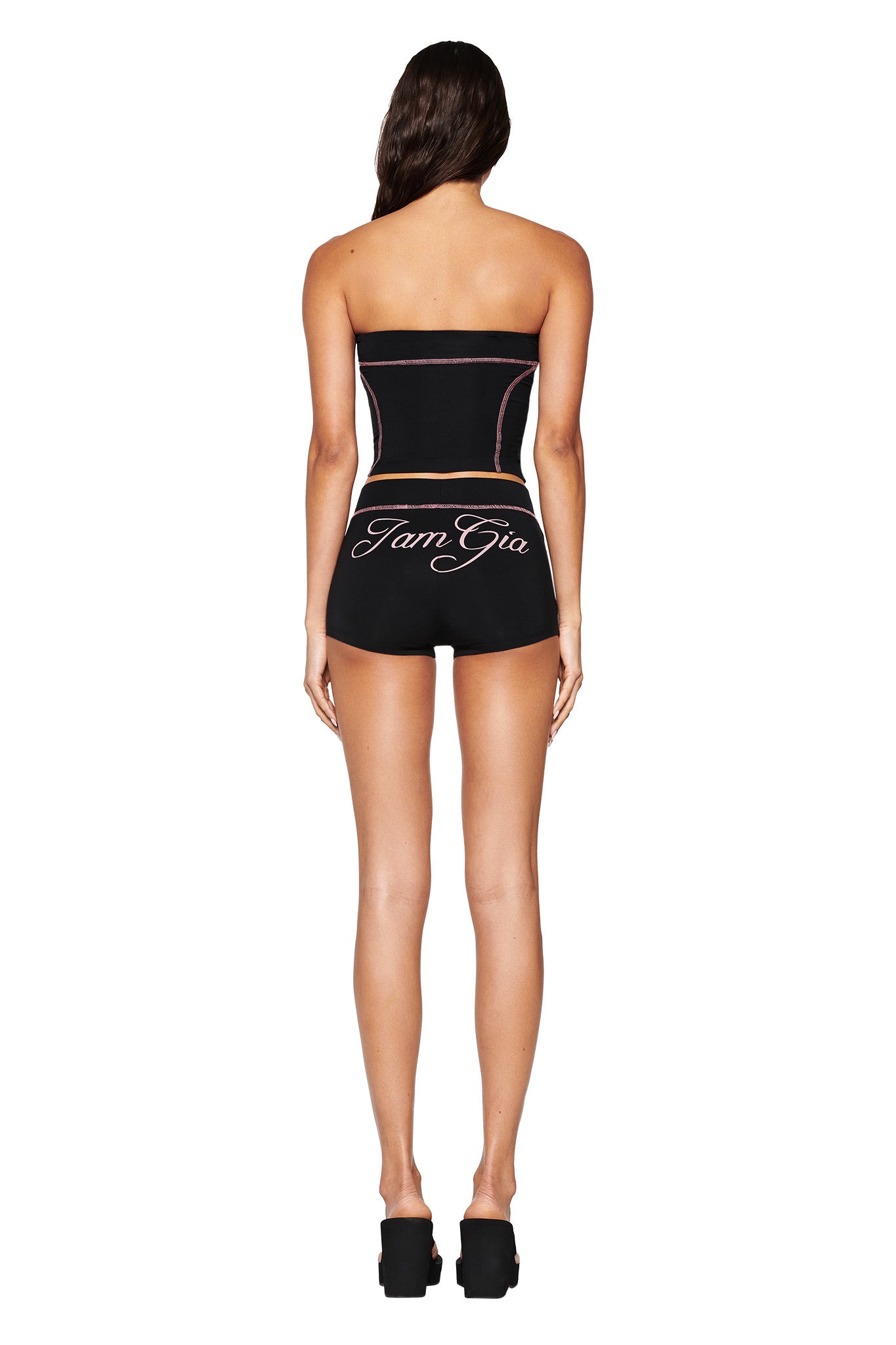 A woman with long hair poses against a white background, wearing I.AM.GIA's black strapless top and the BLARE SHORT - BLACK in a bodycon fit, paired with black platform shoes.