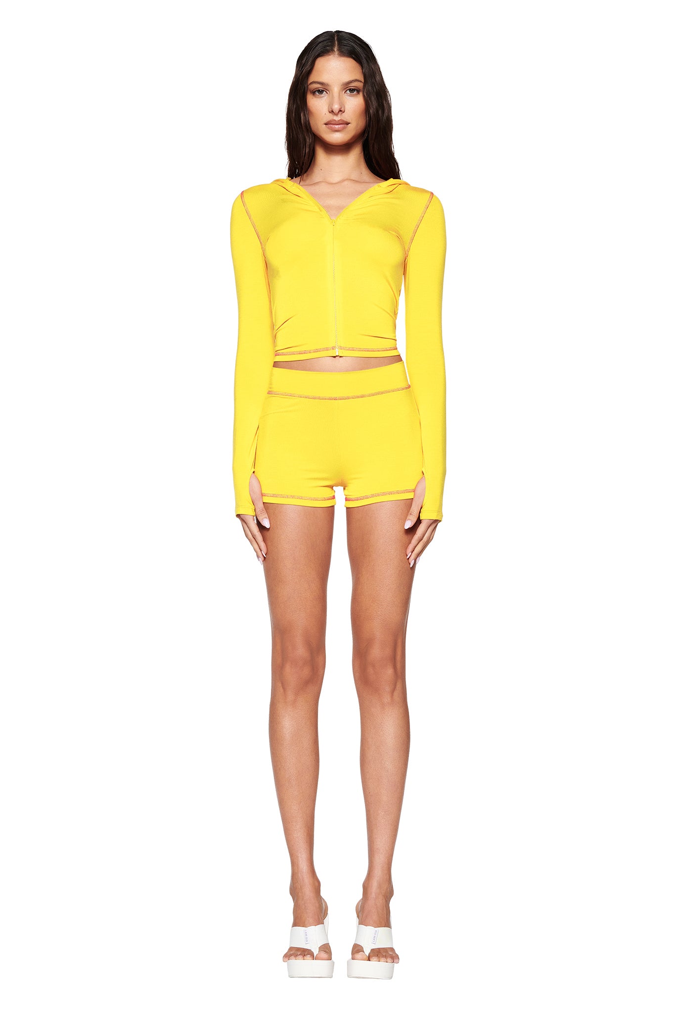 A person poses in a vibrant lemon yellow I.AM.GIA BLARE HOODIE and matching bodycon shorts, paired with white platform sandals against a plain white background.
