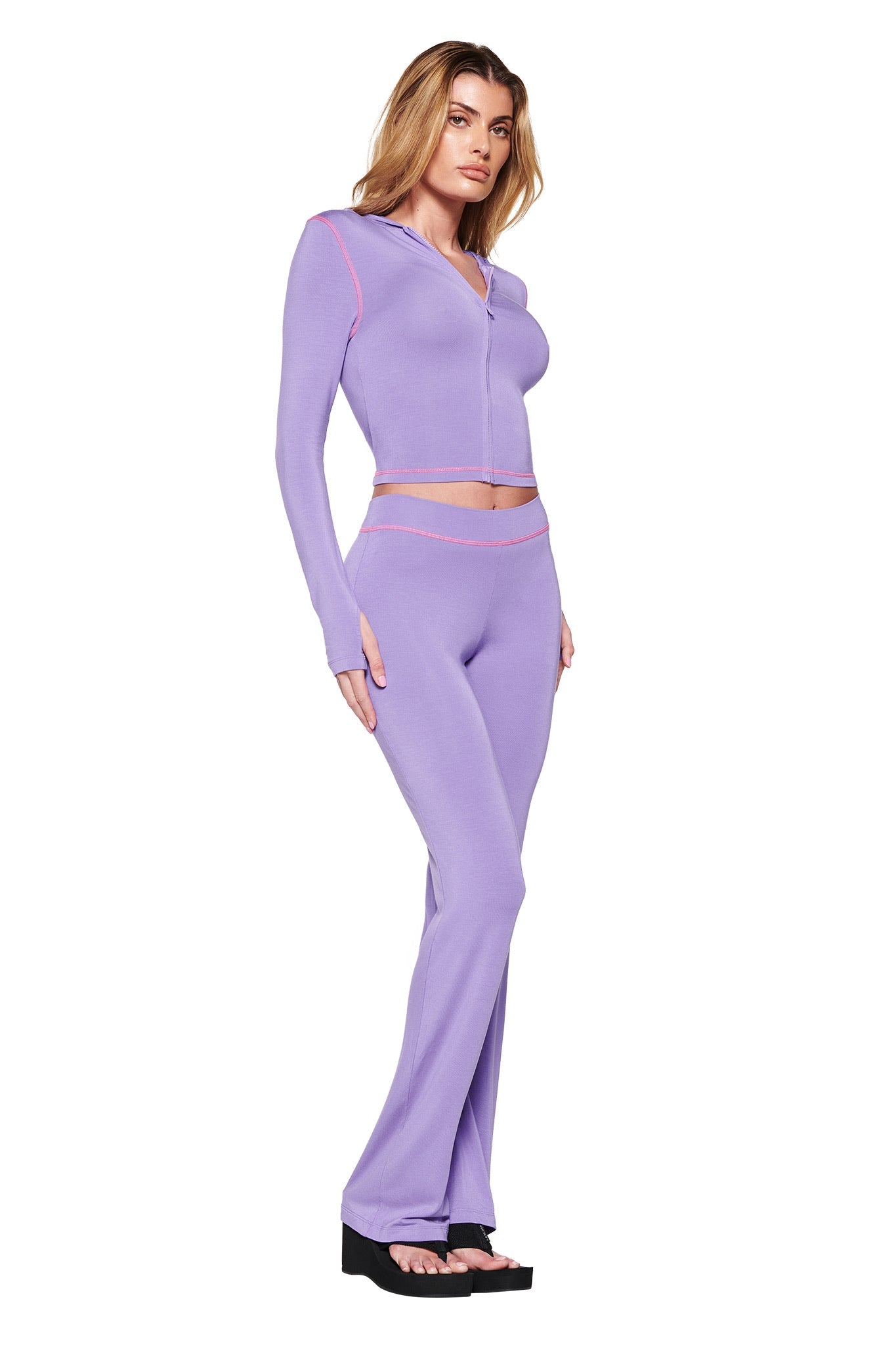 A woman showcases the I.AM.GIA BLARE TRACKPANT - PURPLE, featuring pink trim and a bodycon style. The set includes a zip-up long-sleeve top and matching low-rise pants. She stands slightly to the right in black platform sandals against a plain white background.