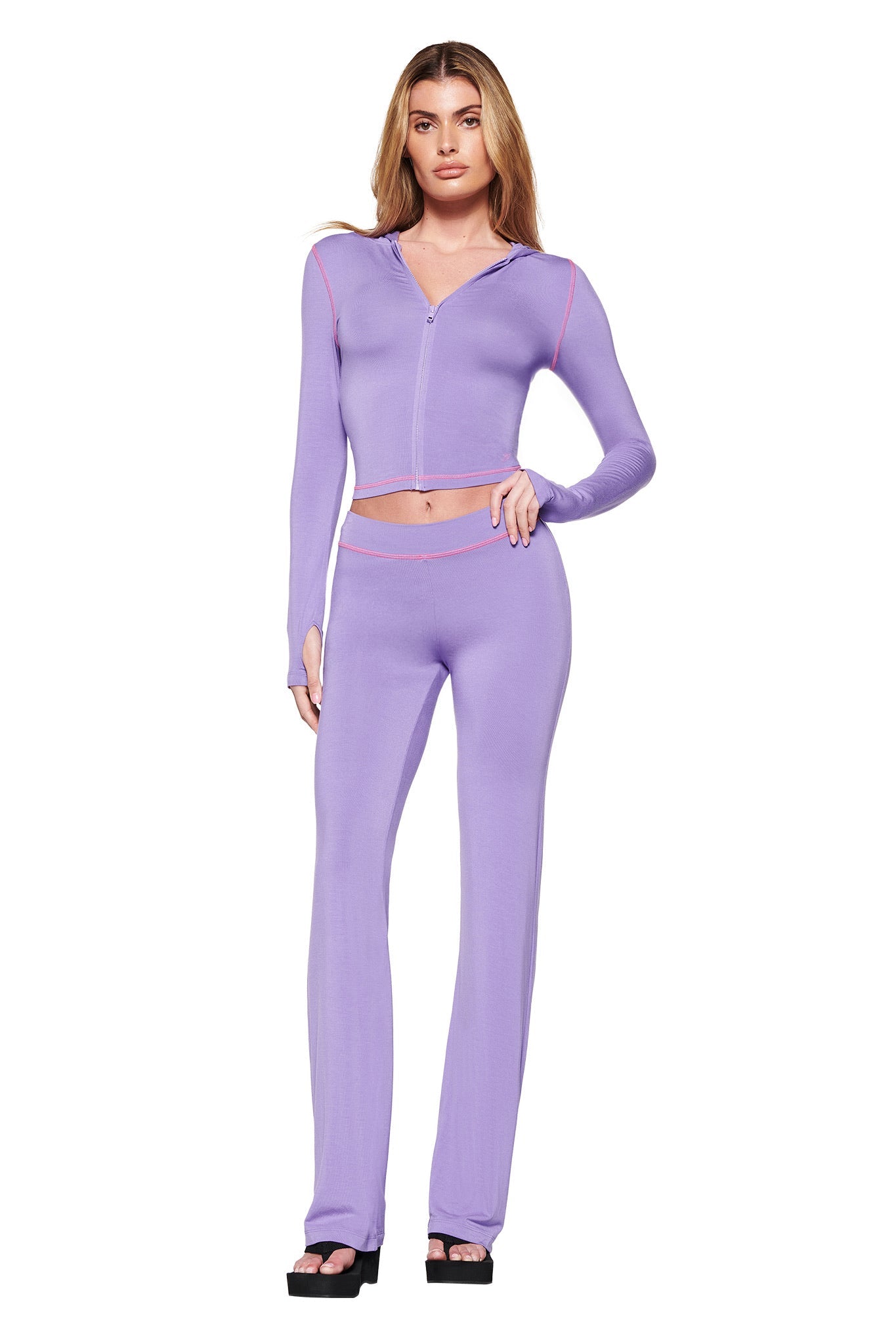 A person models the BLARE HOODIE - PURPLE from I.AM.GIA, showcasing its bodycon fit made from stretchy bamboo fabric. The ensemble features high-waisted pants and black platform sandals, set against a simple white backdrop.