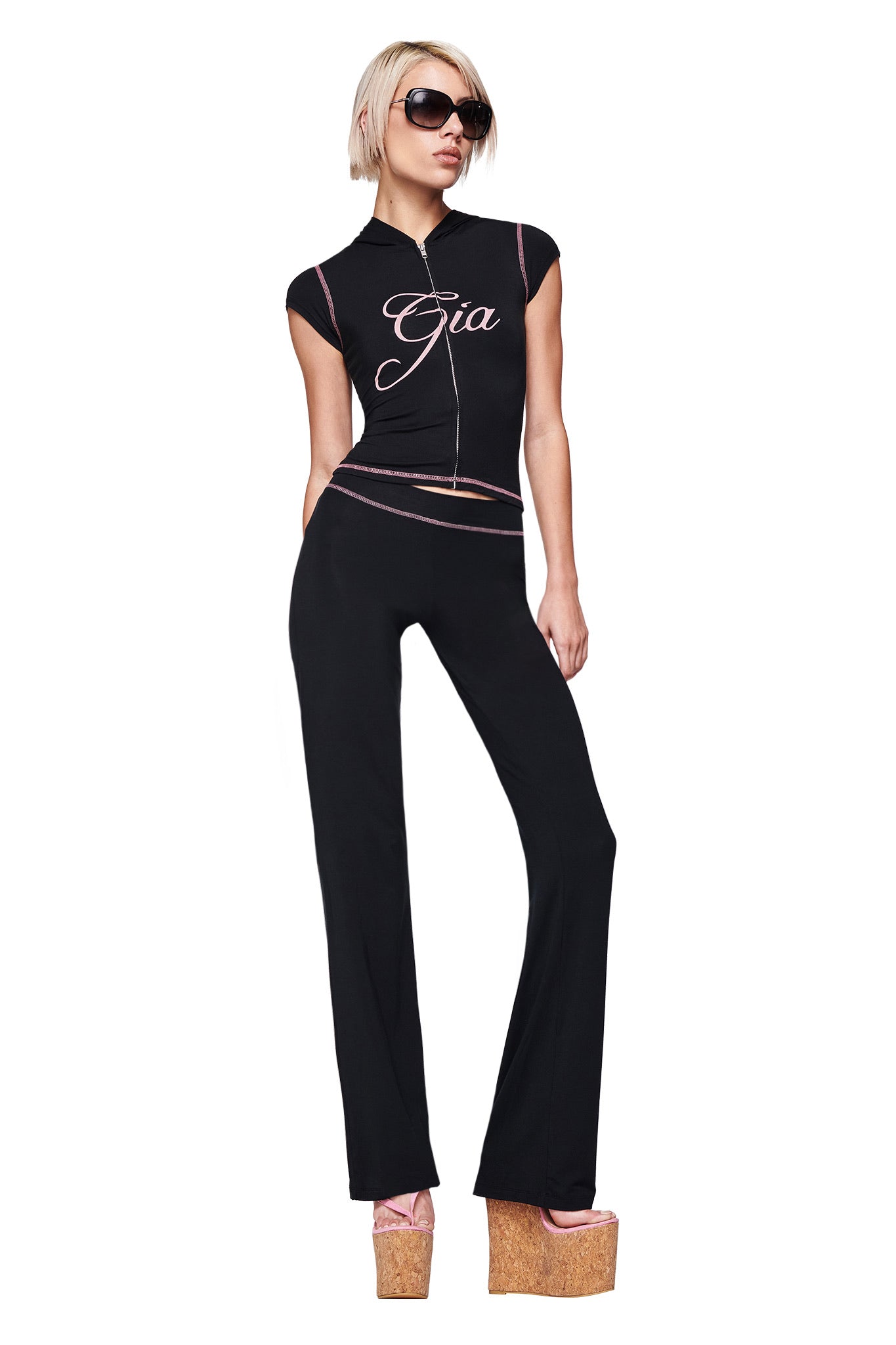 A person with short blonde hair and sunglasses wears the I.AM.GIA BLARE CAP SLEEVE HOODIE in black, paired with black pants featuring pink trim and platform sandals, posing confidently against a white background.