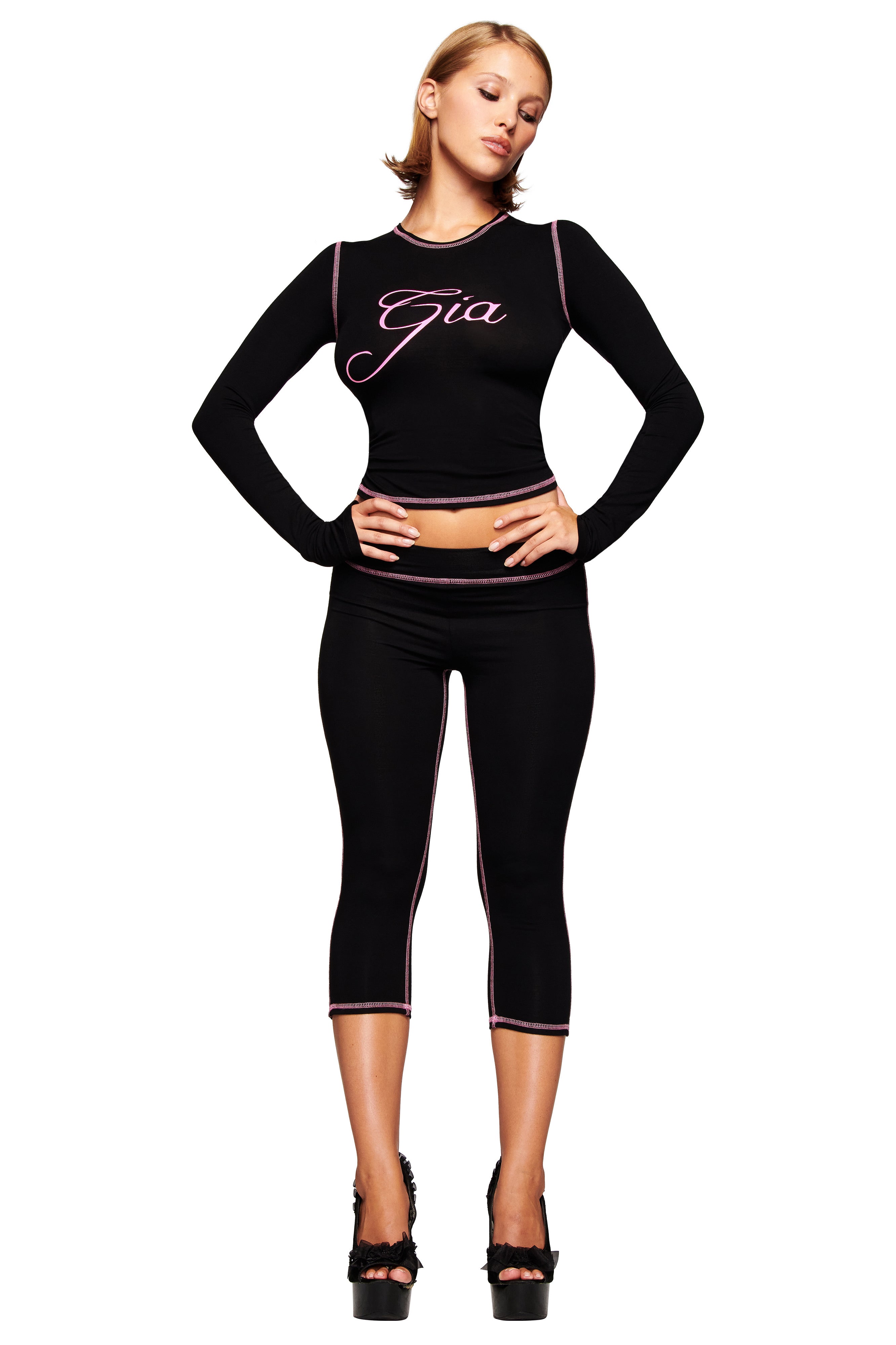 A woman poses against a white background in a fitted black I.AM.GIA long-sleeve crop top, BLARE CAPRI - BLACK leggings with pink piping, and black high-heeled sandals.