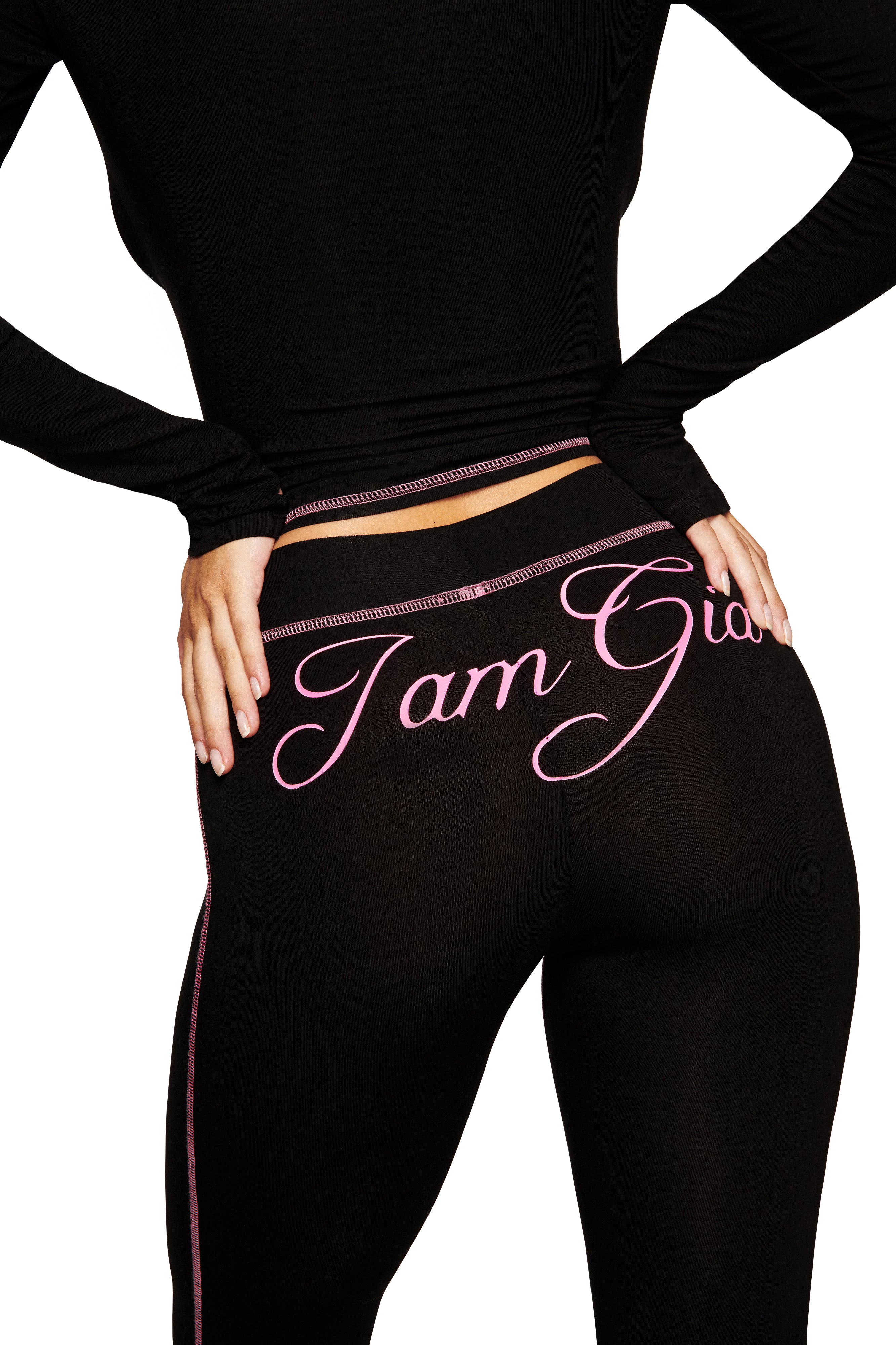 Wearing the BLARE CAPRI - BLACK by I.AM.GIA, with a bold pink I.AM.GIA logo across the back, a person poses confidently in bodycon pants and a long-sleeve top, spotlighting the signature capri style.