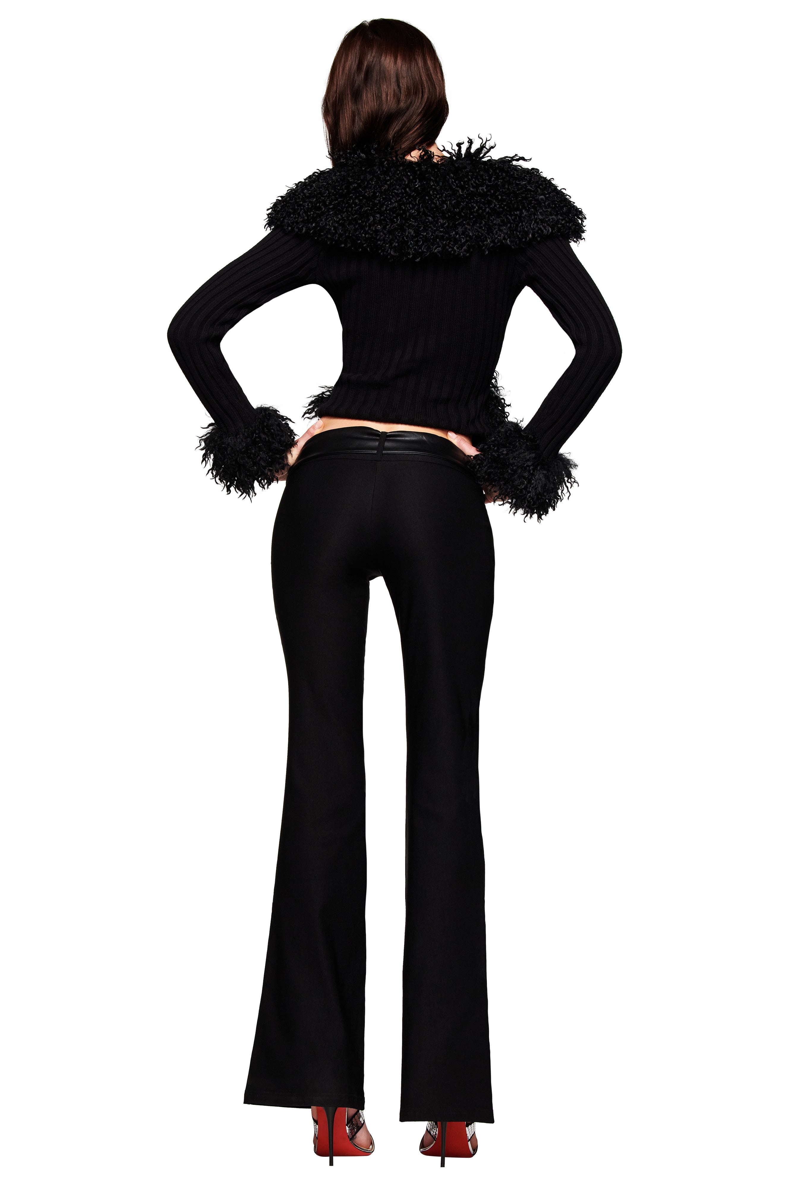 A person stands facing away, wearing black flared pants, red high heels, and the I.AM.GIA CRESSIDA JACKET in black with ribbed knit, shaggy cuffs, and a faux fur collar.