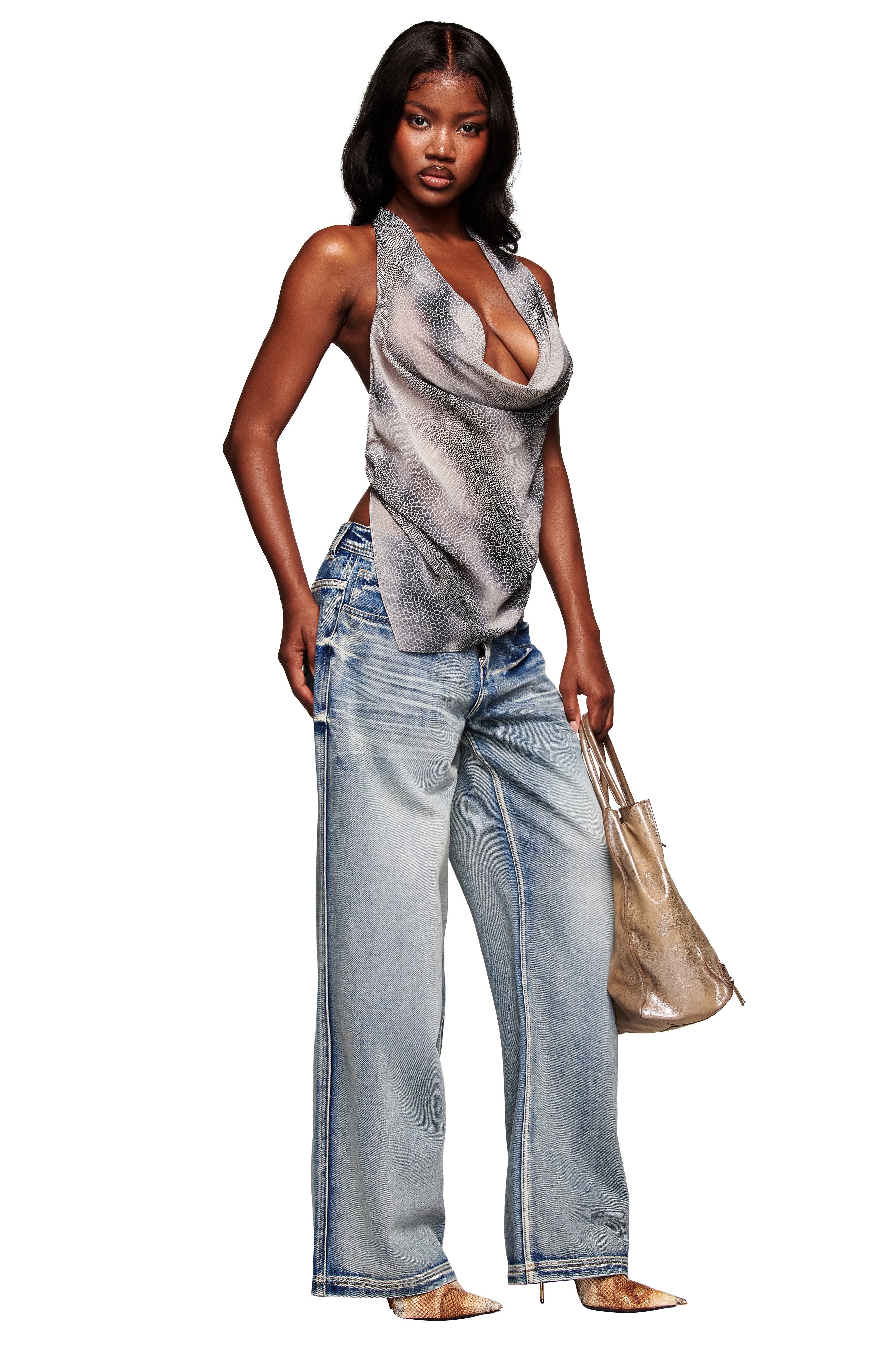 A woman wears the I.AM.GIA PALISADE JEAN in blue, paired with a sleeveless draped top and metallic beige tote. She stands confidently in pointed heels against a white background.