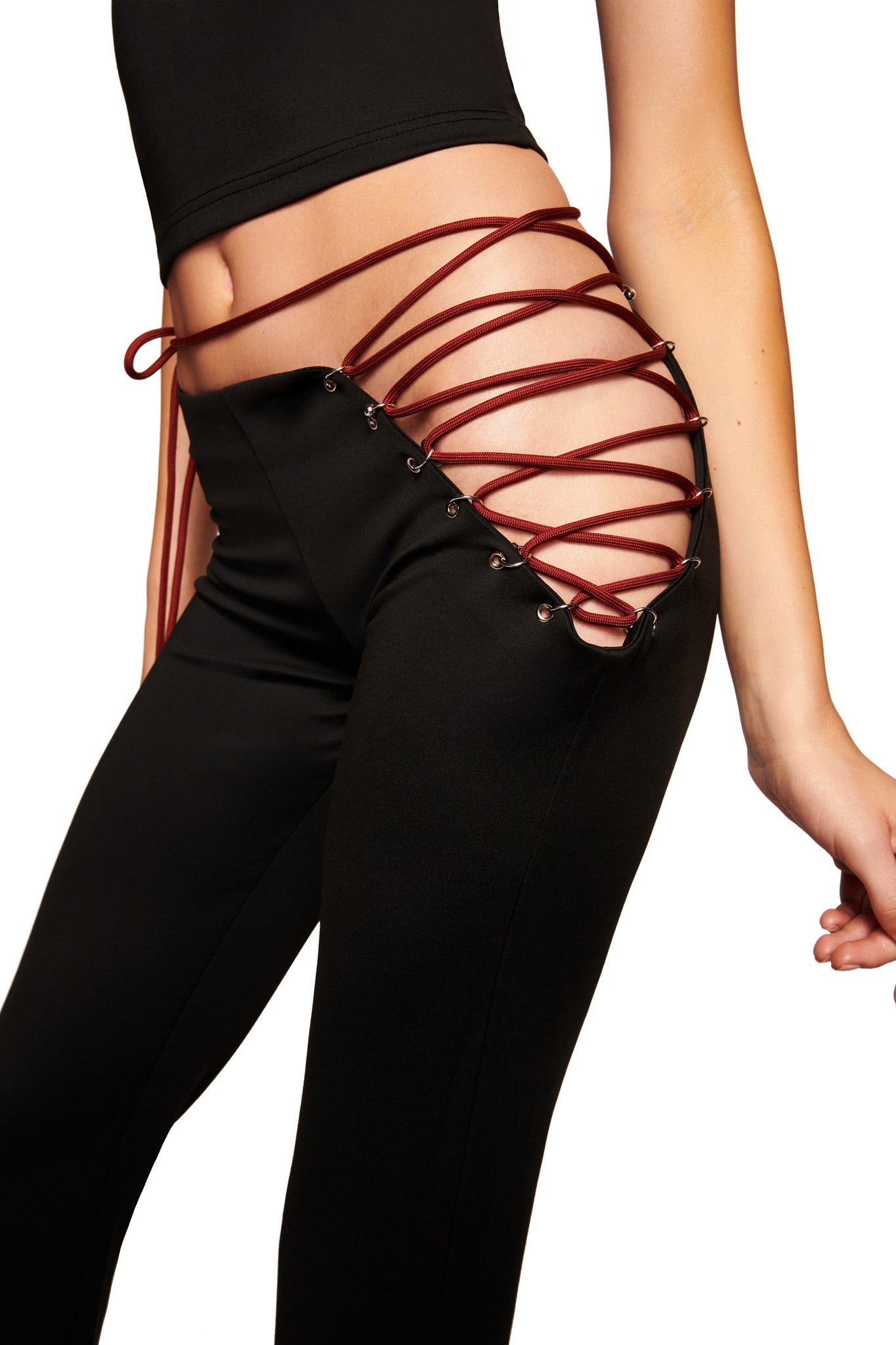 Wearing the I.AM.GIA ONYX PANT - BLACK, which features a low rise fit, exposed hip cutout, and crisscross red lace-up detail with metal eyelets, paired with a sleek black top and hand resting by their side.