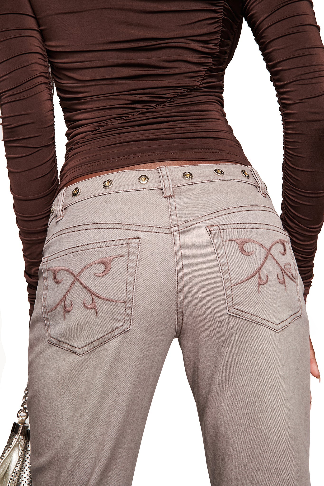 The image shows the back waist-down view of a person wearing I.AM.GIA’s GREER PANT - GREY, featuring a V-shaped waistband and back pocket stitching, paired with a fitted ruched brown top and holding a small handbag.
