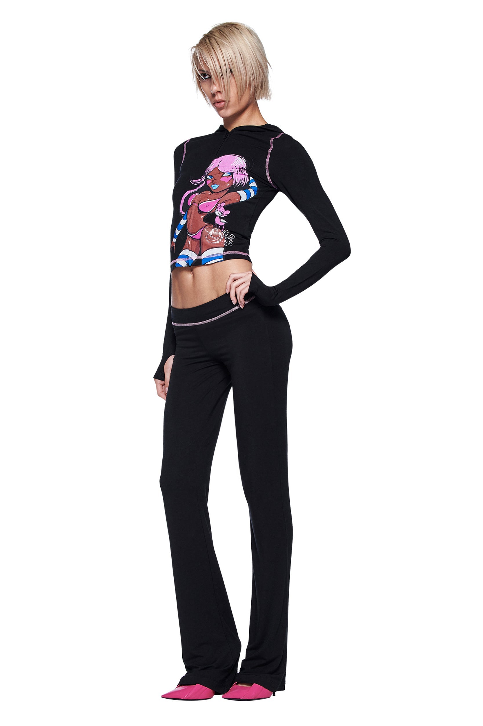A person with short blond hair wears a fitted black long-sleeve top with a colorful character, I.AM.GIA's FAFI BLARE PANT in black, and pink shoes, standing against a white background.