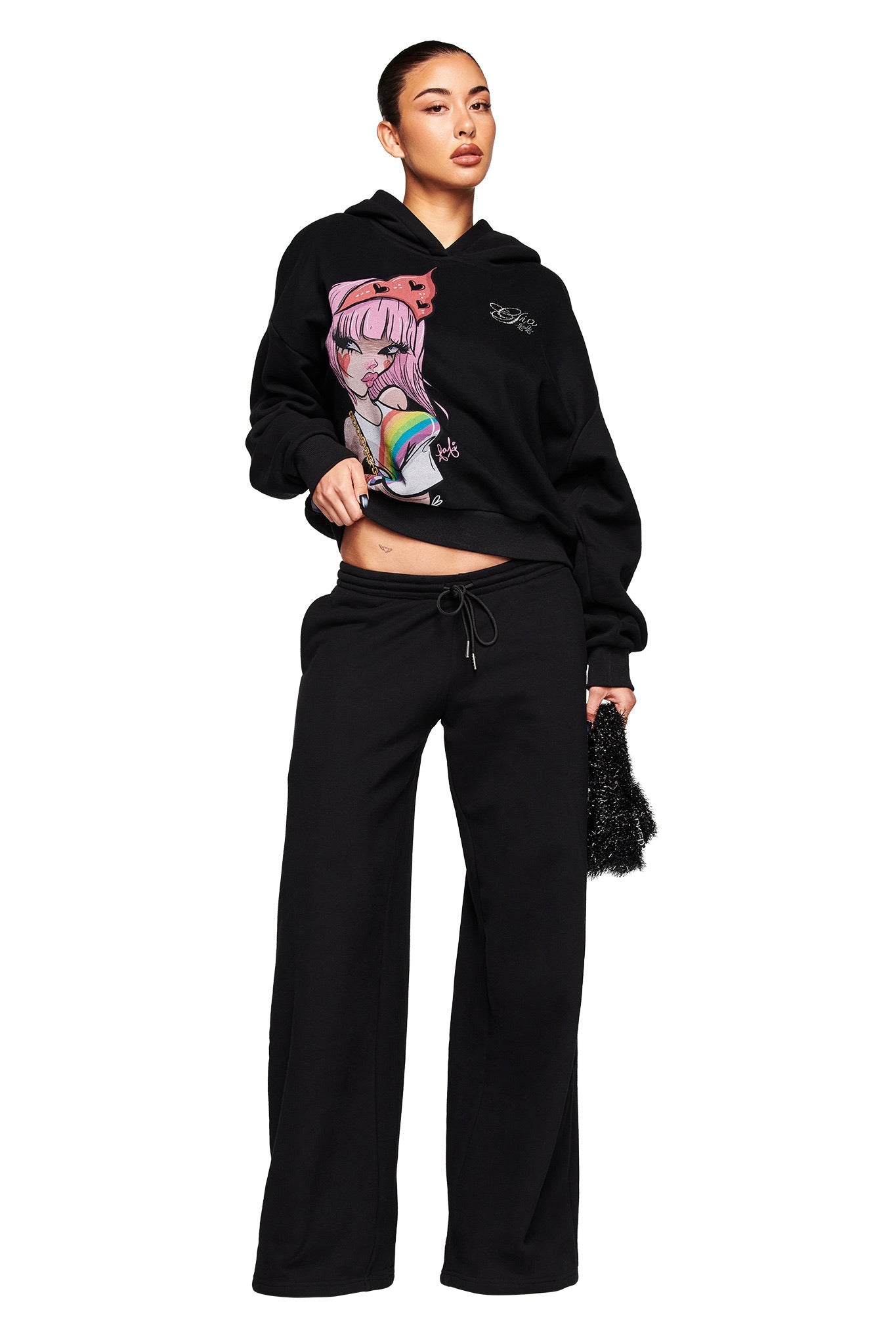 A woman wears the I.AM.GIA FAFI TRACKPANT in black and a black hoodie with a vibrant anime-style rainbow graphic, holding a small textured black handbag. Her hair is styled in a sleek bun.