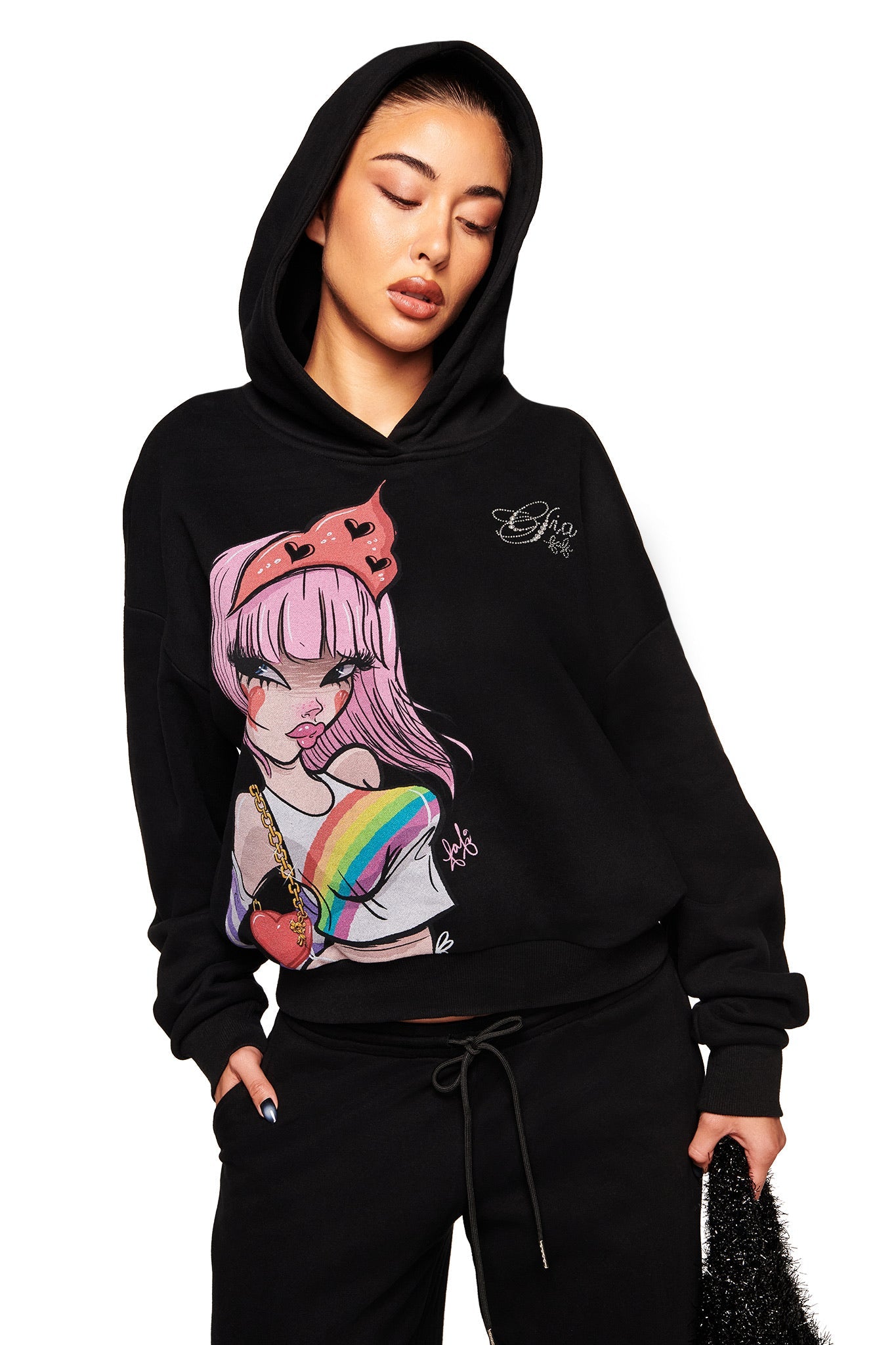 A woman wears the I.AM.GIA FAFI HOODIE - BLACK, featuring a colorful anime-style Fafinettes girl graphic, paired with black drawstring pants. She stands with eyes closed and one hand in her pocket.