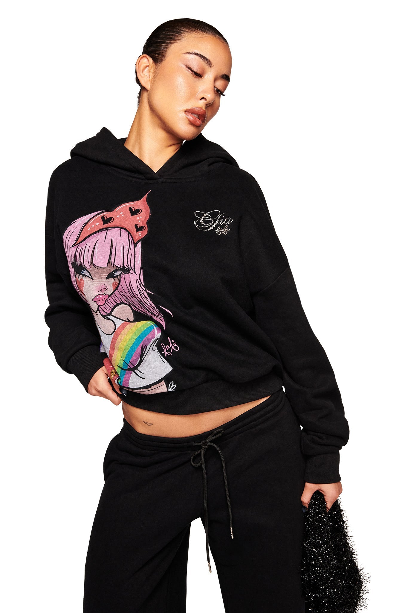 A woman poses in the I.AM.GIA FAFI HOODIE - BLACK, an oversized hoodie with a colorful Fafinettes anime-style graphic, paired with black pants and a textured black handbag against a white background.