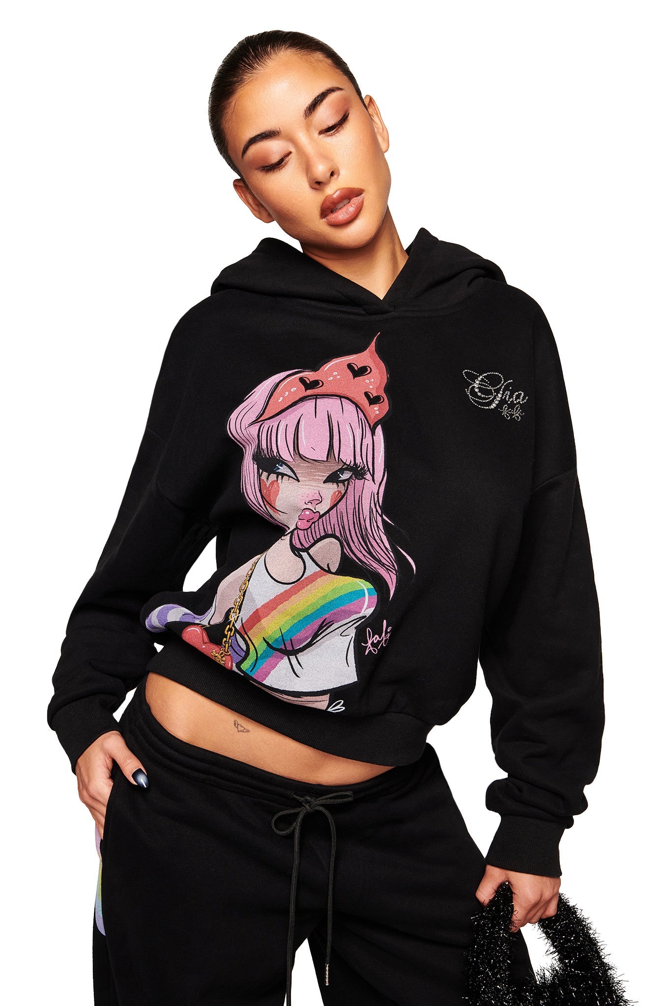 A woman stands with her eyes closed and head tilted, wearing the I.AM.GIA FAFI HOODIE - BLACK, an oversized hoodie featuring vibrant anime-inspired art of a pink-haired, red-crowned girl inspired by Fafinettes.